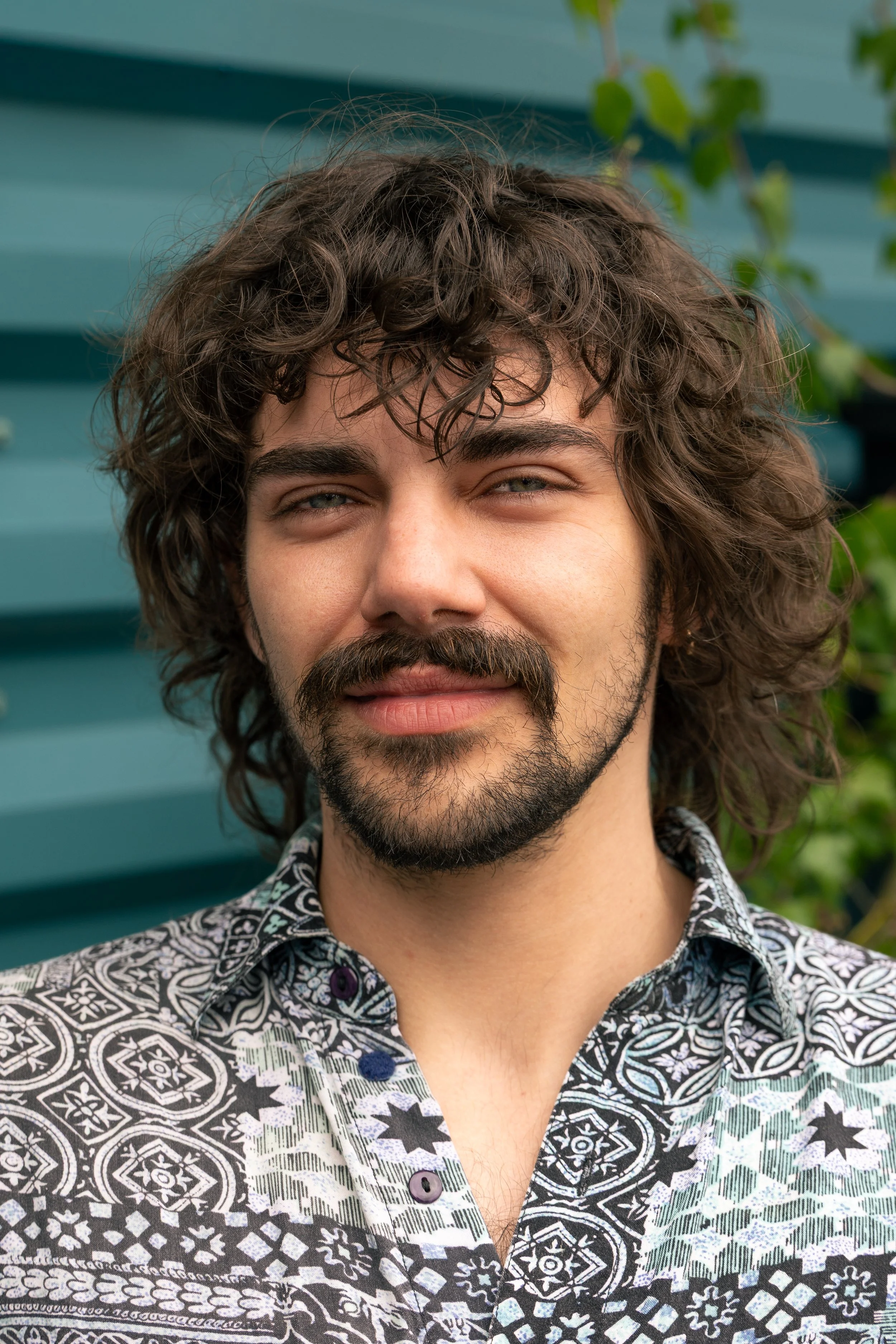 A man with curly brown hair and a beard, smiling, wearing a patterned black and white shirt with purple buttons, standing outdoors in front of a teal-colored structure and green foliage.