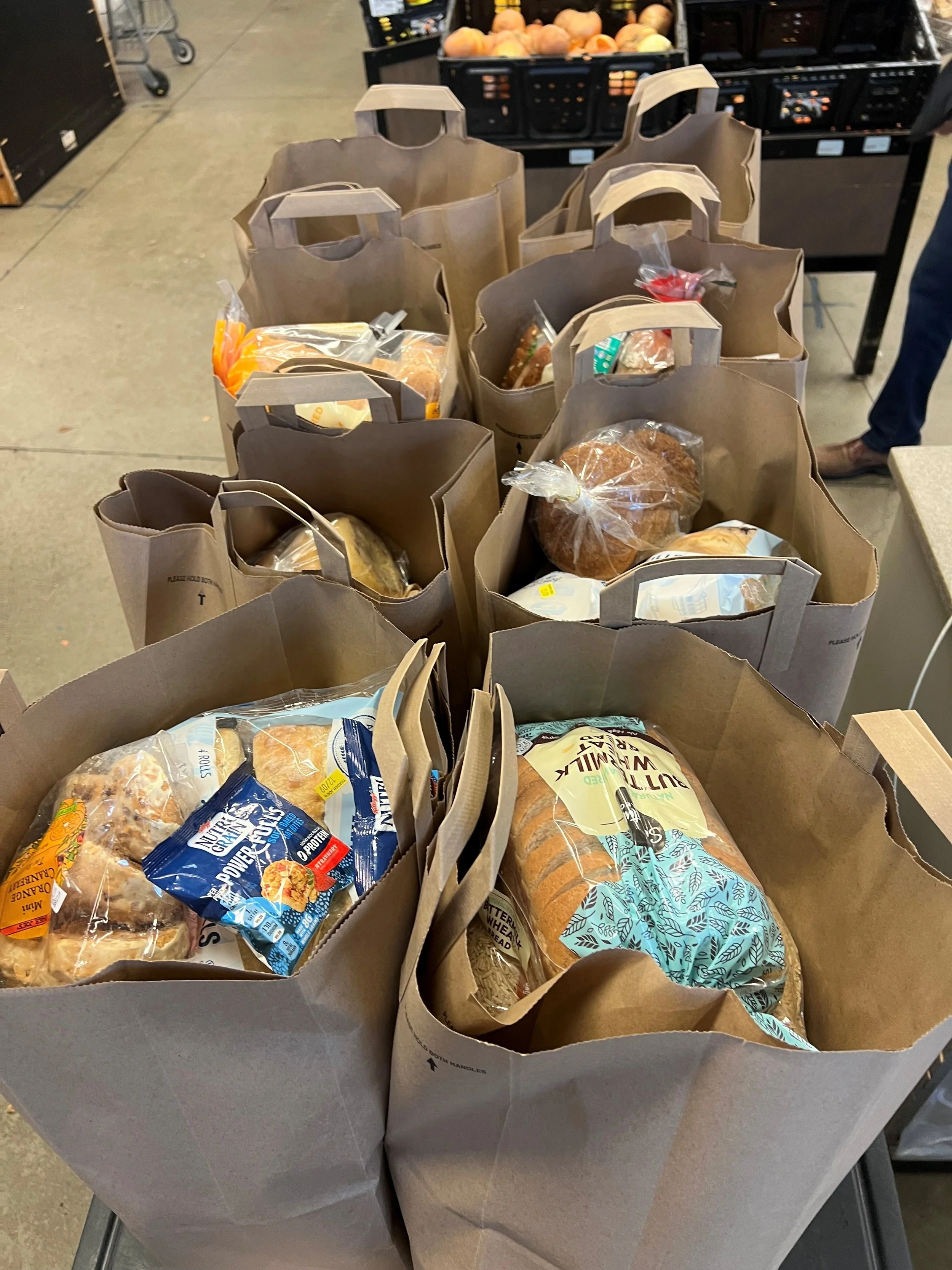 Multiple paper grocery bags filled with various types of bread and baked goods on a shopping cart in a grocery store.
