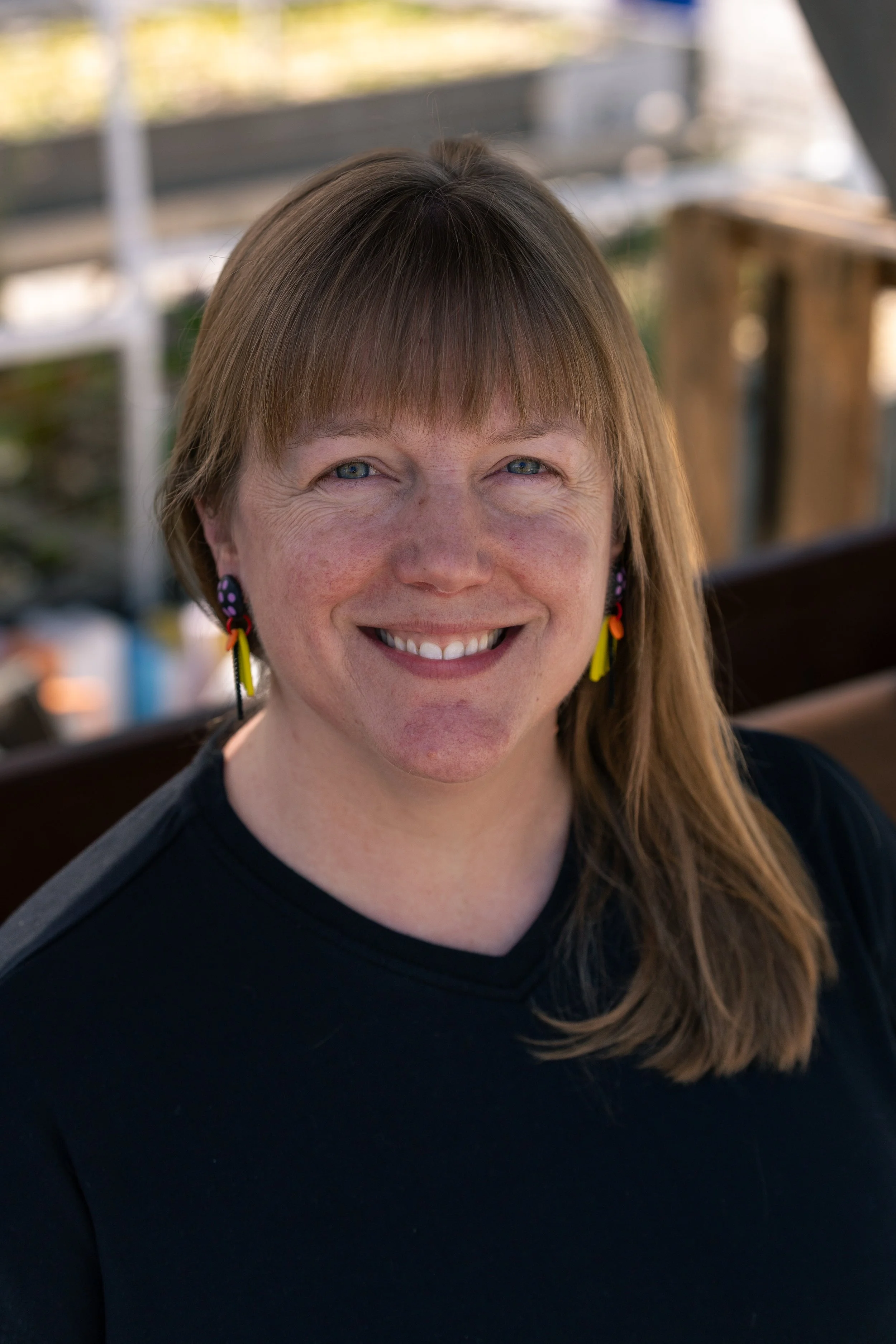 A woman with light skin, light brown hair with bangs, wearing colorful earrings, smiling, outdoors with a blurred background of a bridge and trees.