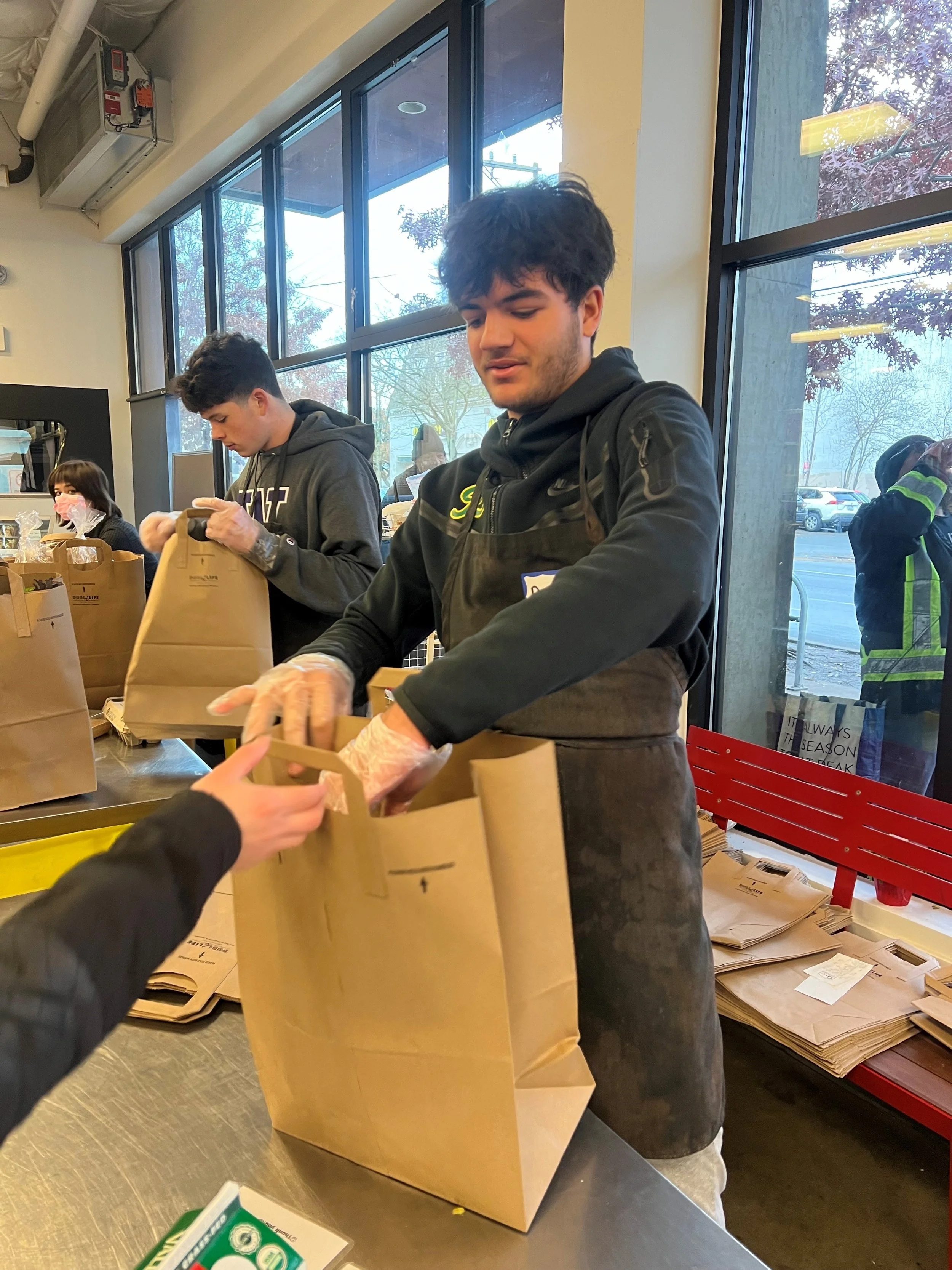 People packing brown paper bags at a counter inside a store, with large windows showing parked cars and trees outside.
