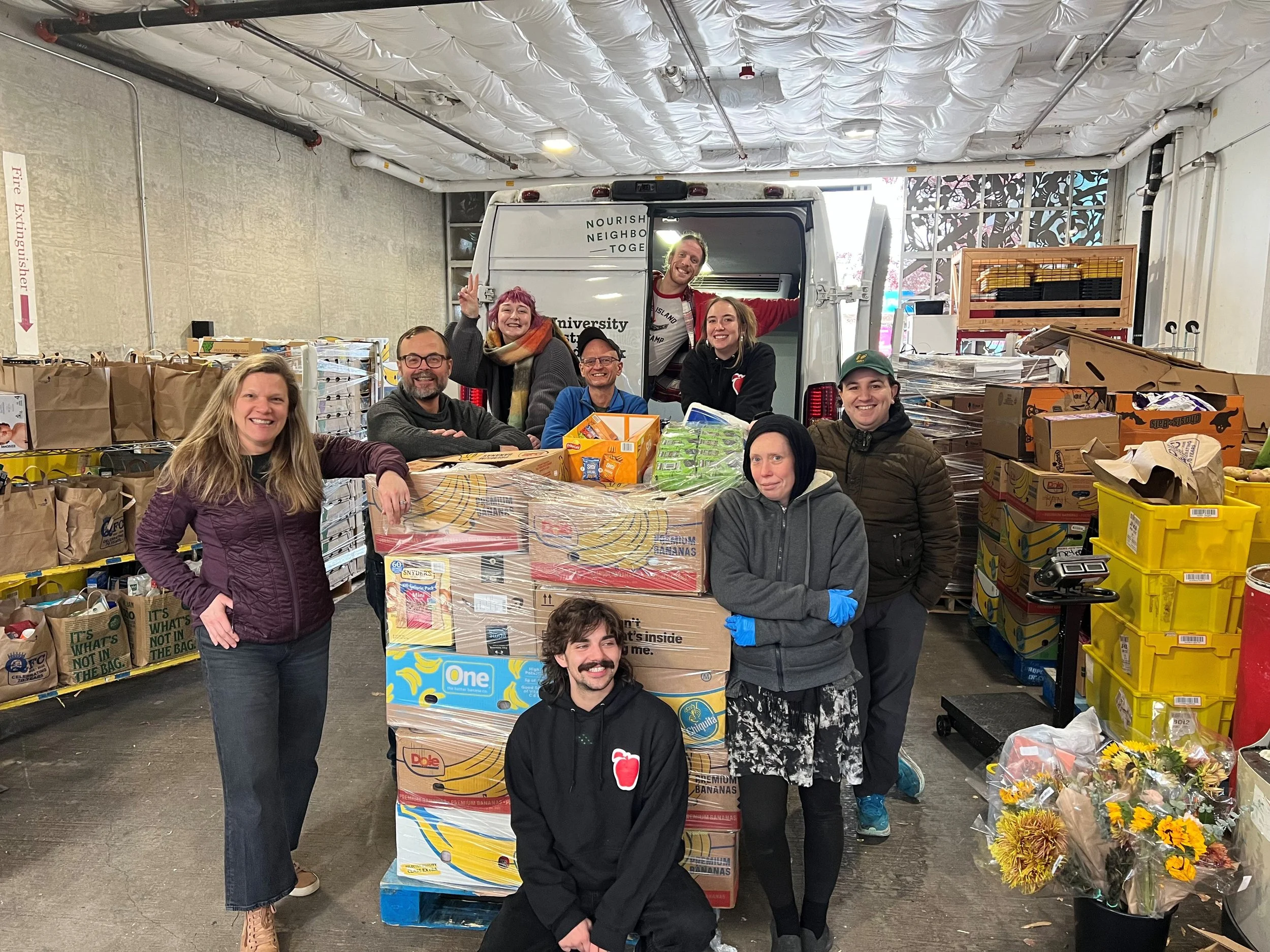 Group of people smiling and posing inside a warehouse with shelves filled with boxes and packages, standing near a truck.