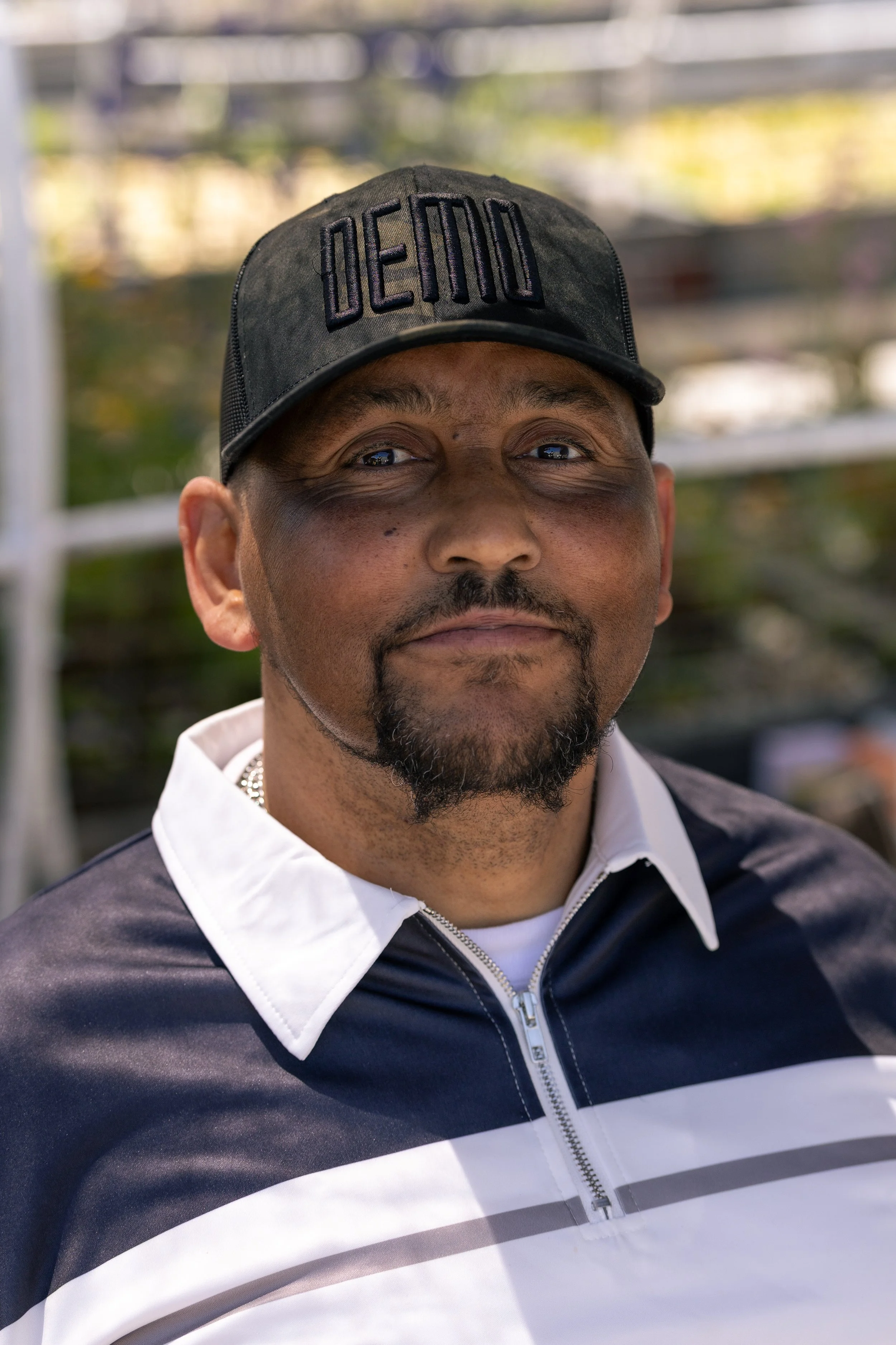 Close-up of a man wearing a black cap with the word 'DEAD' embroidered in black, smiling slightly, with a blurred outdoor background.