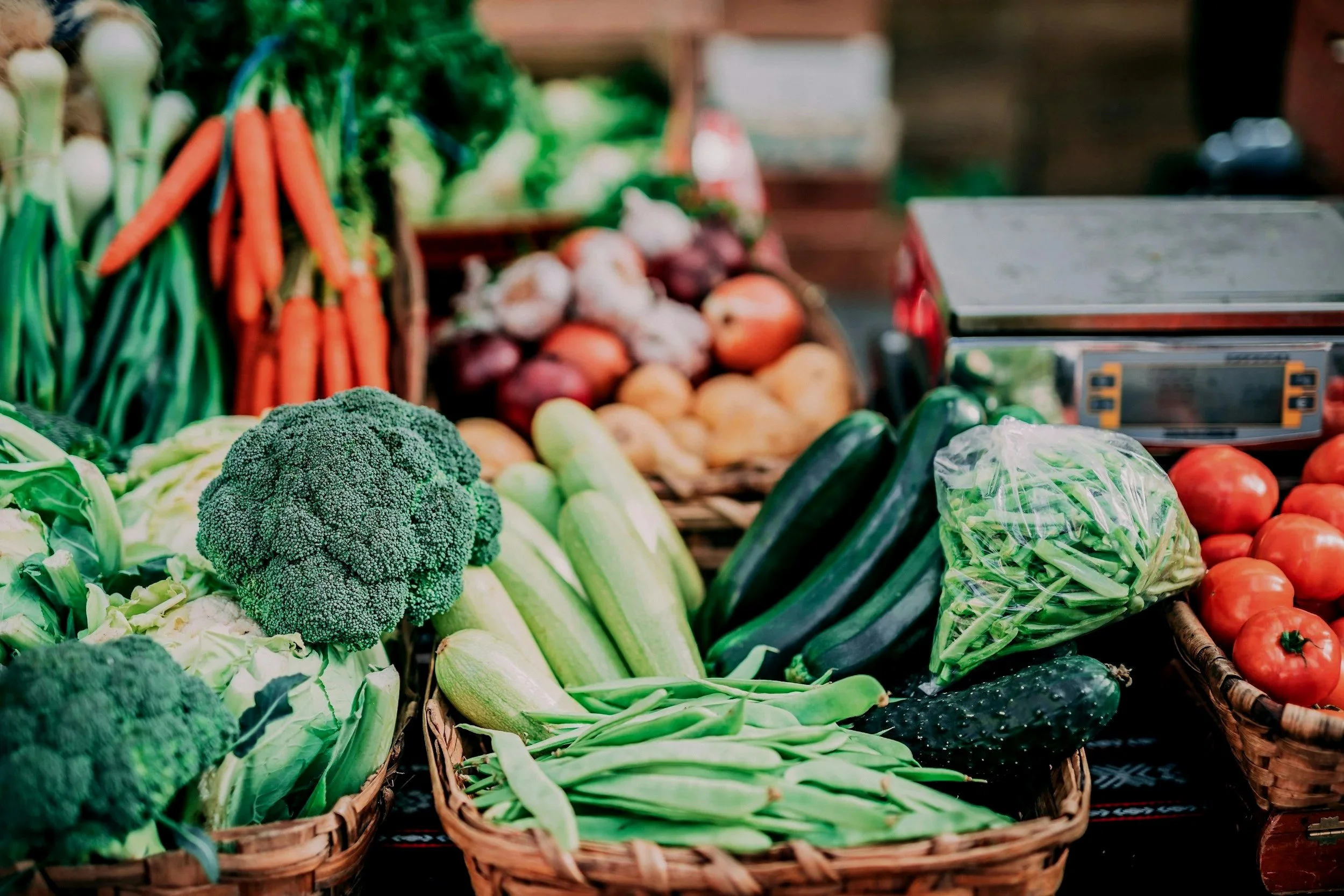 Fresh vegetables at a market, including broccoli, carrots, onions, tomatoes, and cucumbers displayed in baskets.