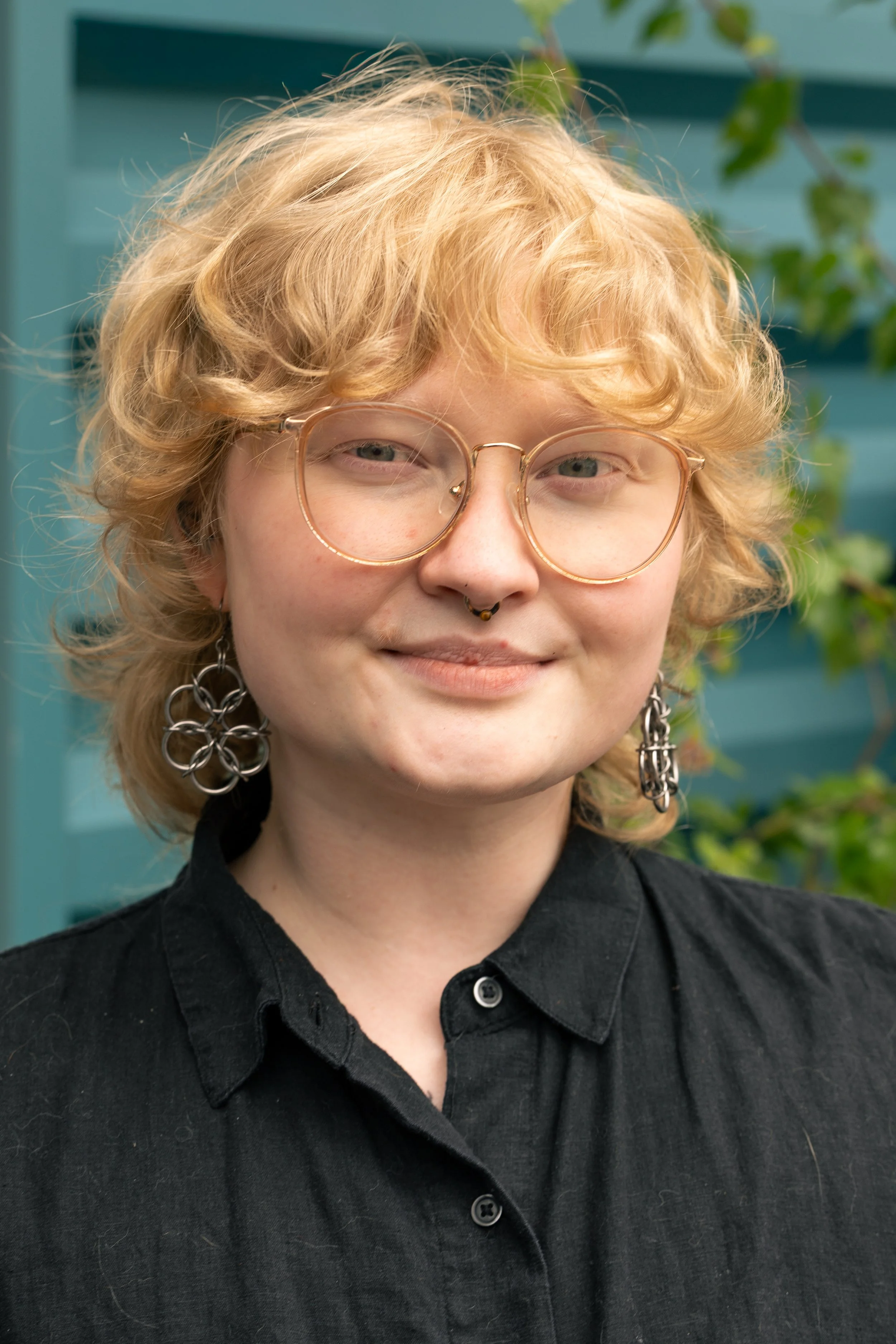 Close-up of a person with blond curly hair, wearing round glasses, a septum piercing, and large earrings, dressed in a black shirt, standing in front of a teal background with green foliage.