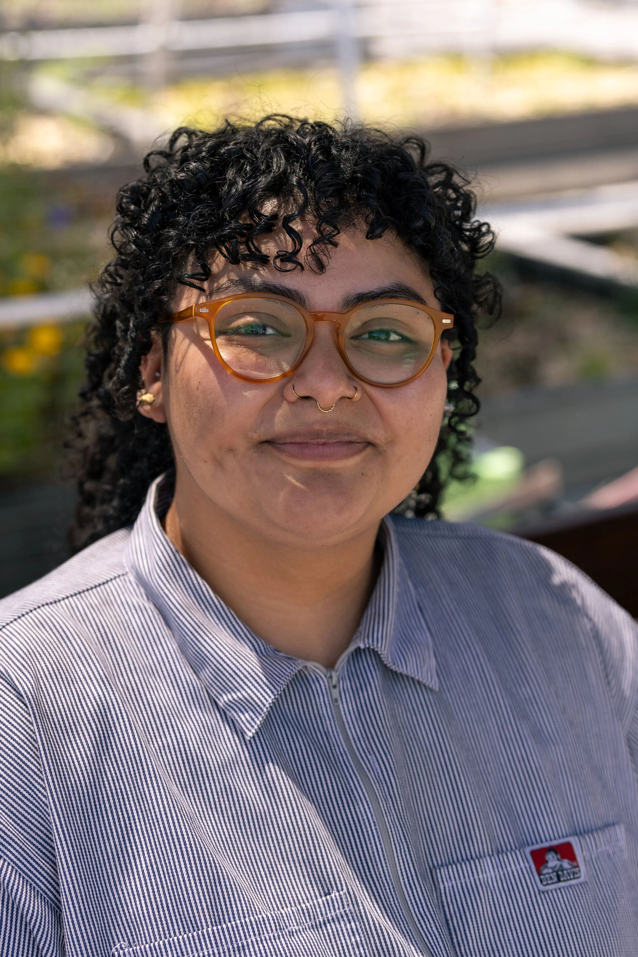 Close-up of a young woman with curly black hair, wearing round orange glasses and a striped button-up shirt with a logo.