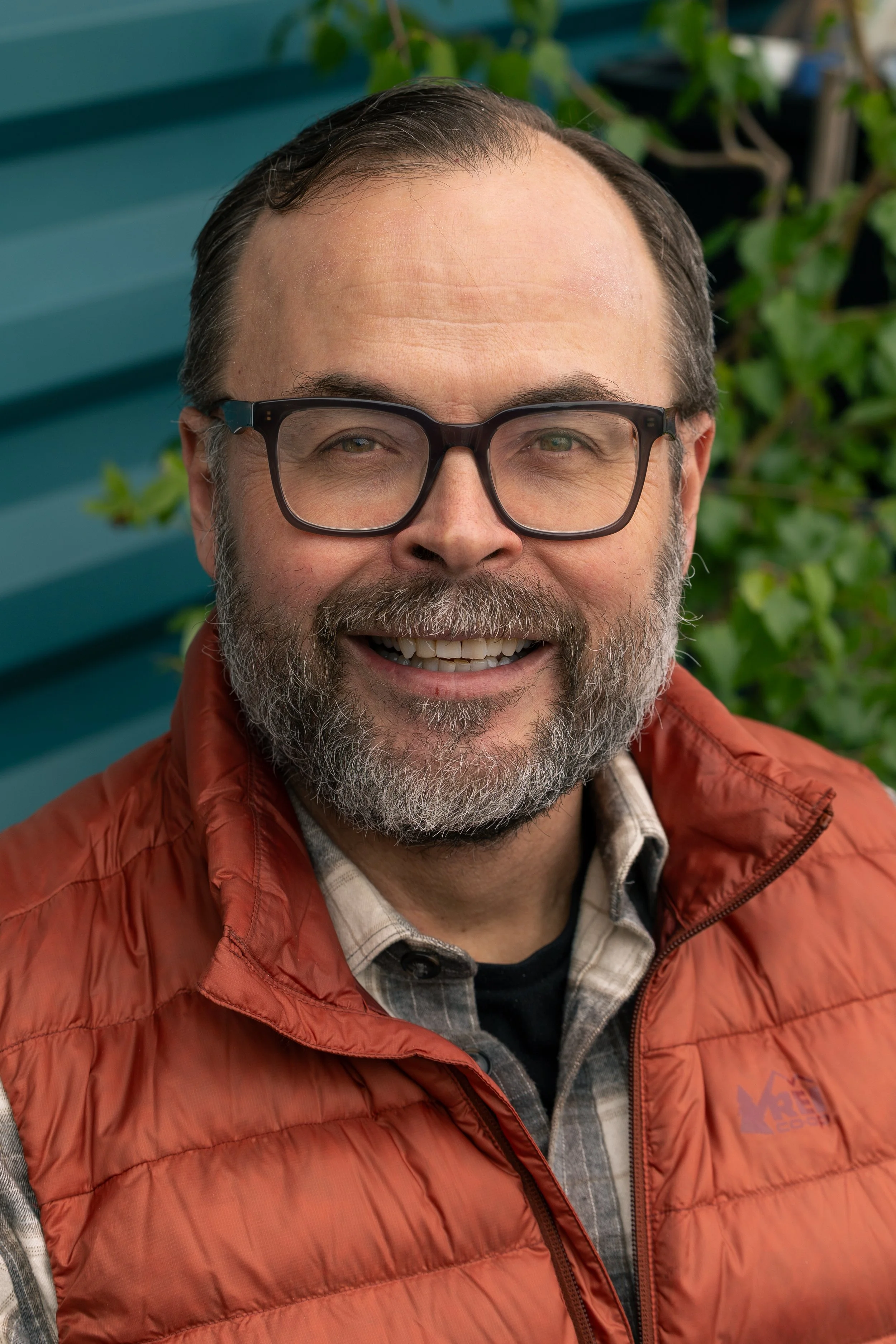 Close-up of a smiling man with glasses, a beard, wearing an orange puffer jacket and a plaid shirt, outdoors with green foliage and a teal-colored structure in the background.