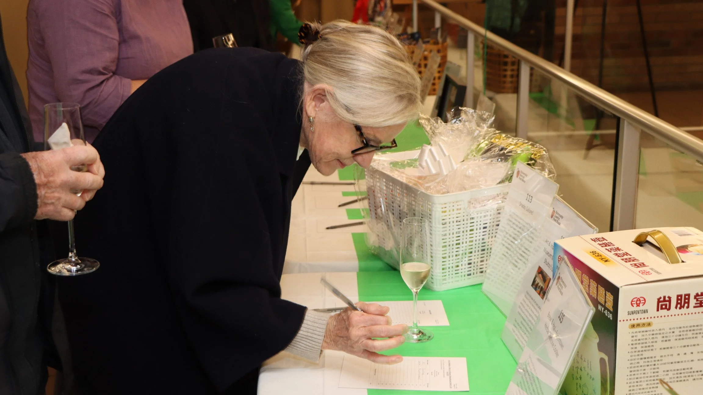 An elderly woman with gray hair and glasses writing on a notepad with a pen at a registration or information table, with a partly filled glass of champagne in front of her, surrounded by informational signs and baskets at an event.