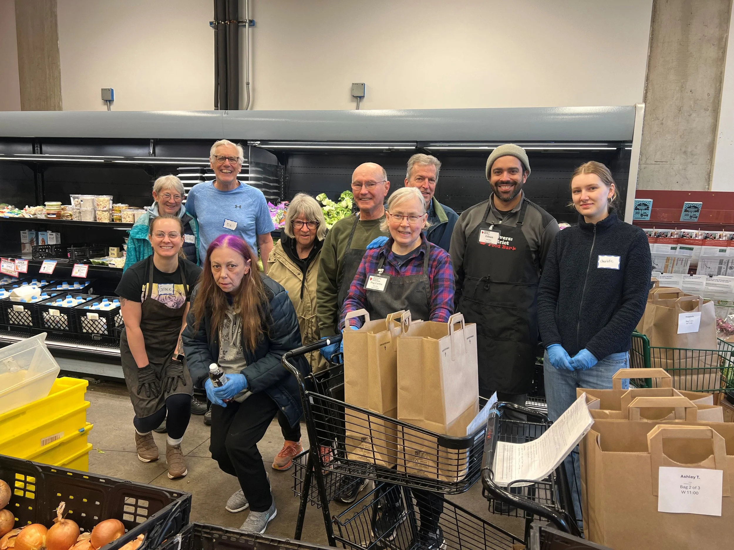 A group of smiling volunteers, diverse in age and ethnicity, shopping for groceries in a store, with bags and shopping carts around them, during a food bank event.