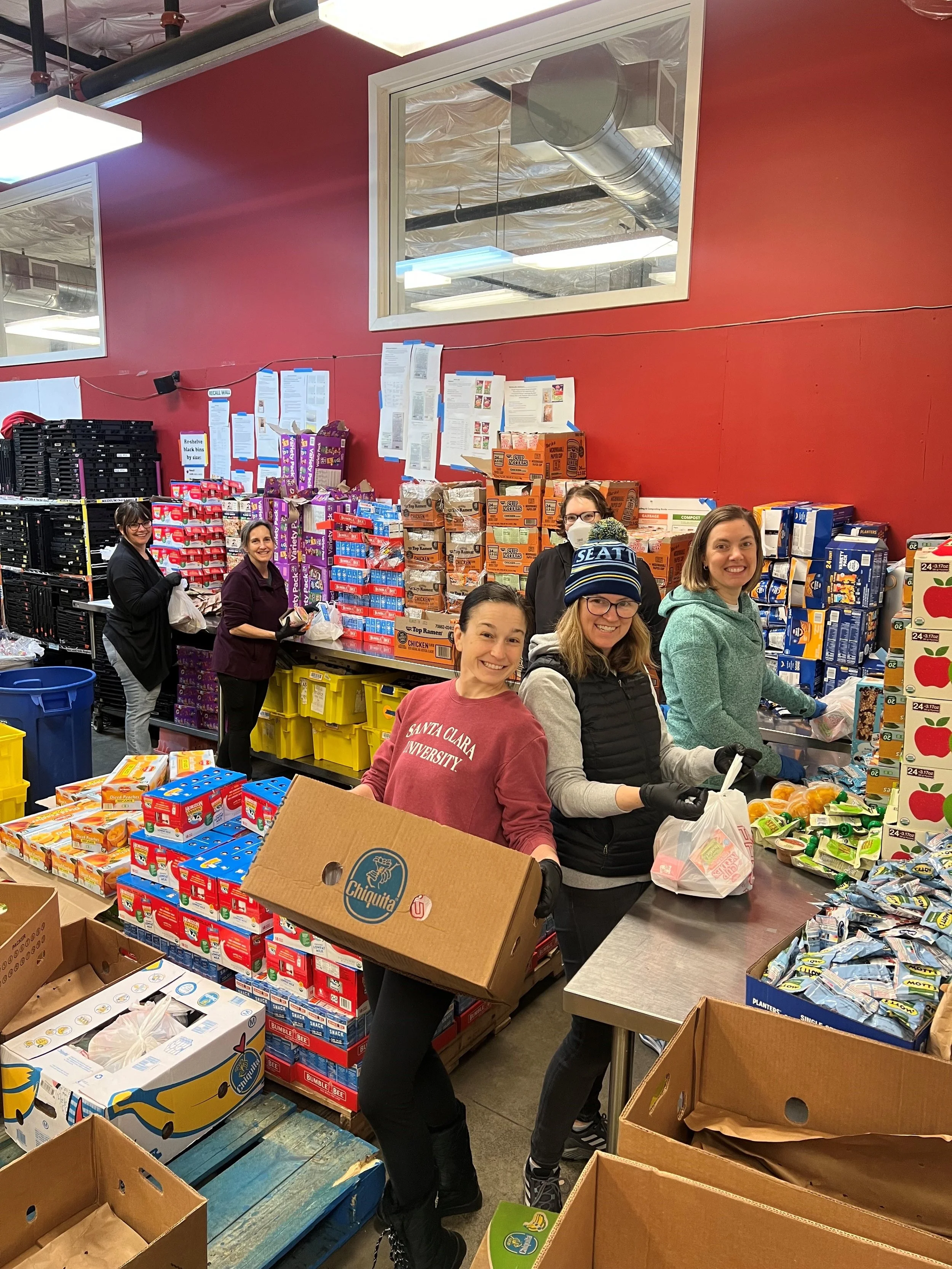 Group of people volunteering at a food donation or packing event in a grocery store, with boxes of snacks and food items on tables and shelves.