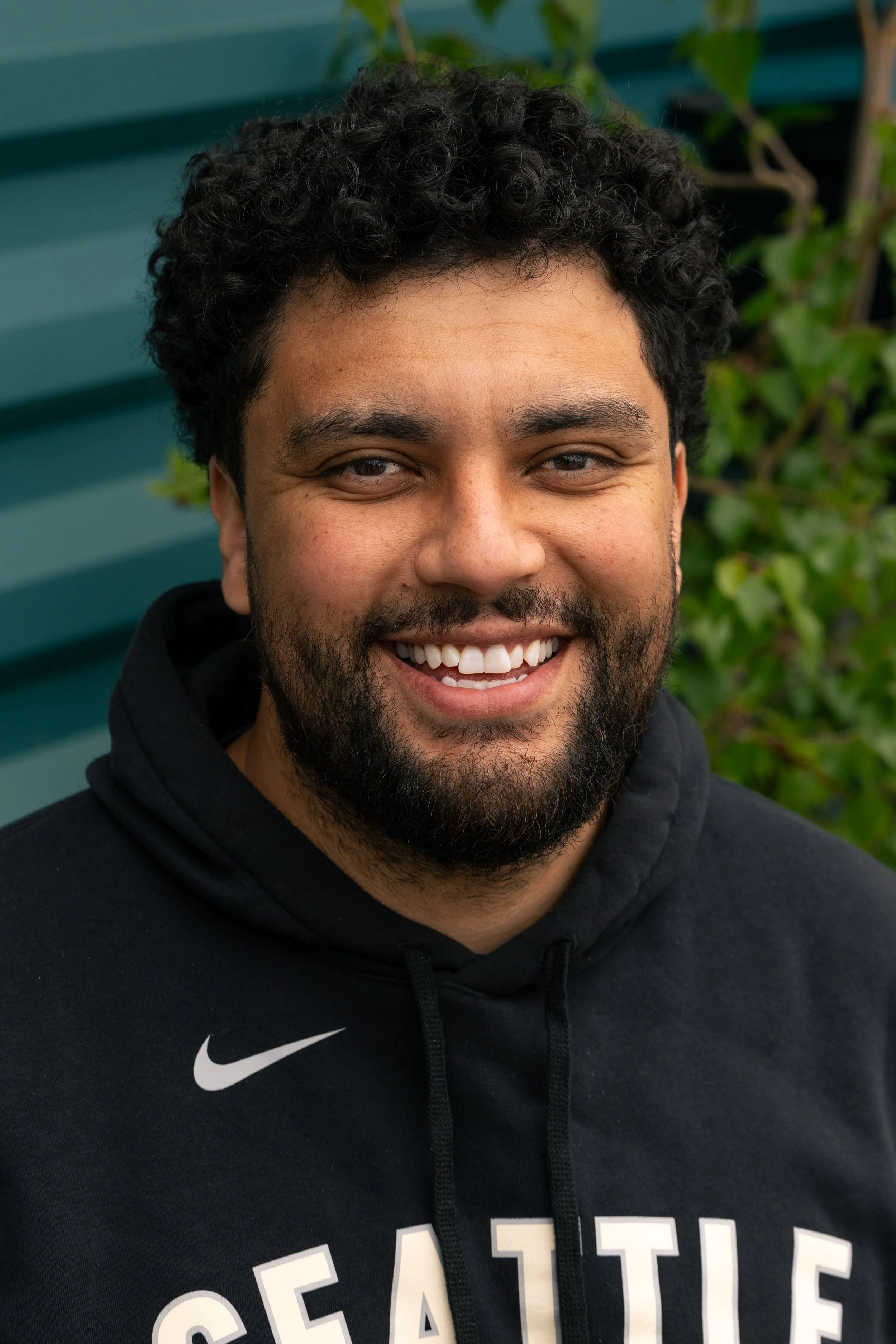 Close-up of a smiling man with curly black hair and a beard, wearing a black Nike hoodie with white text, standing outdoors with green foliage and a teal-colored wall in the background.