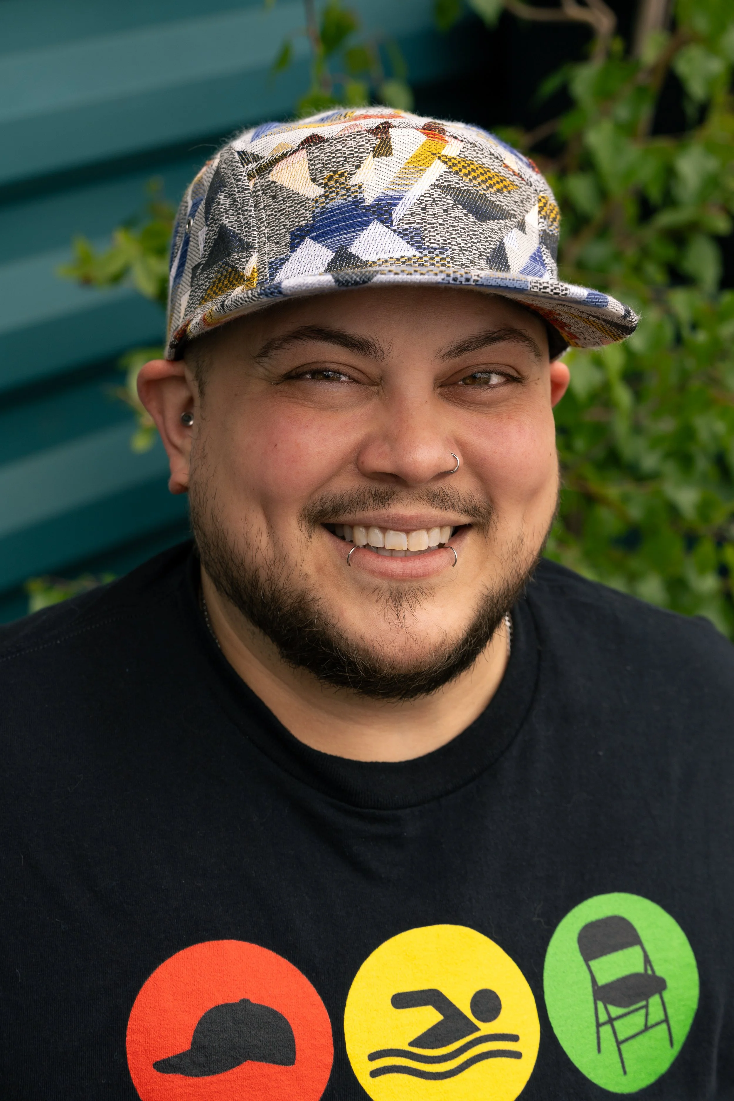 Close-up of a smiling person wearing a patterned cap and a black T-shirt with colorful icons and text, standing outside near a green fence with greenery in the background.