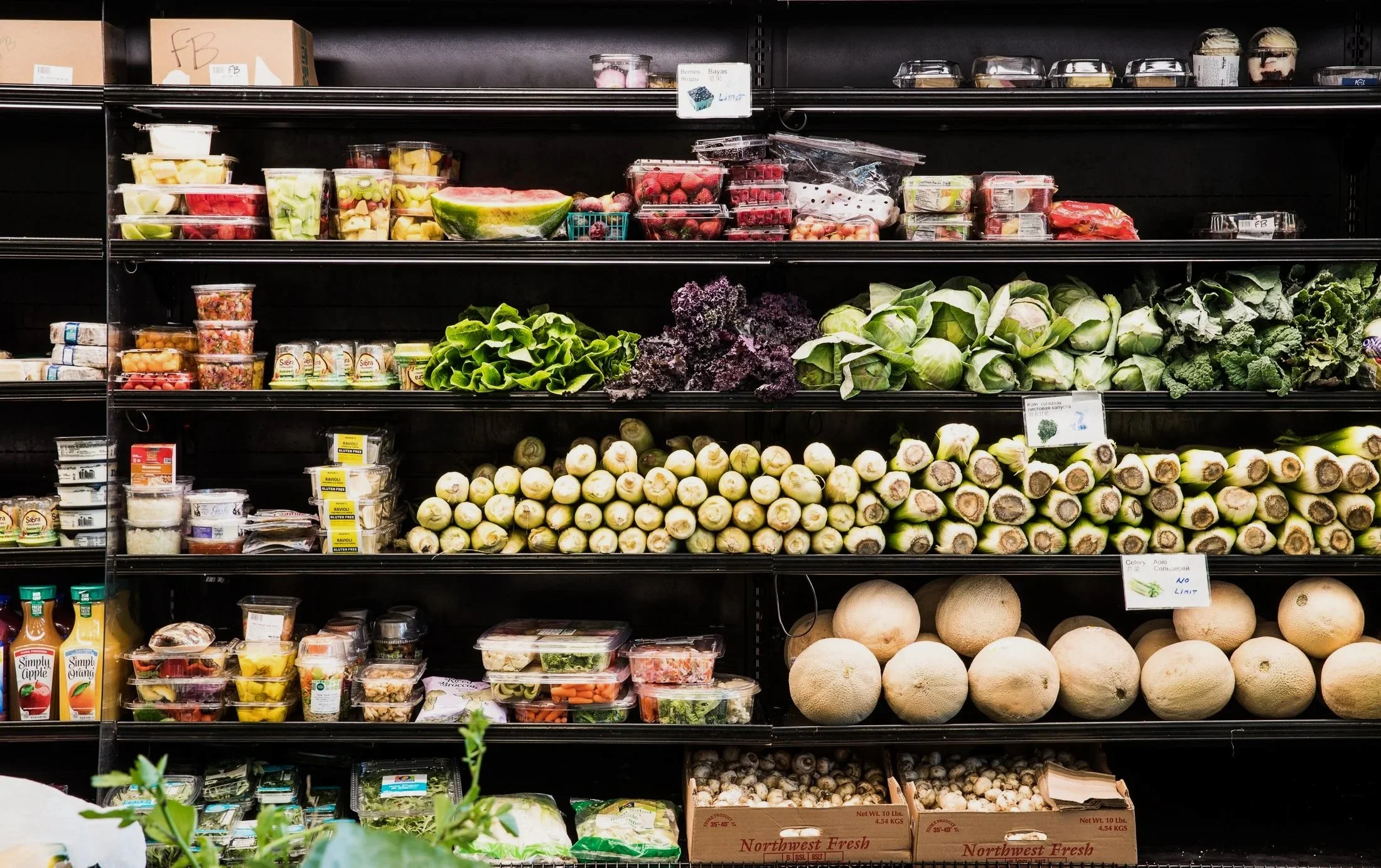 Display of fresh fruits, vegetables, and packaged food items on supermarket shelves, including melons, lettuce, strawberries, blueberries, and pre-packaged salads.