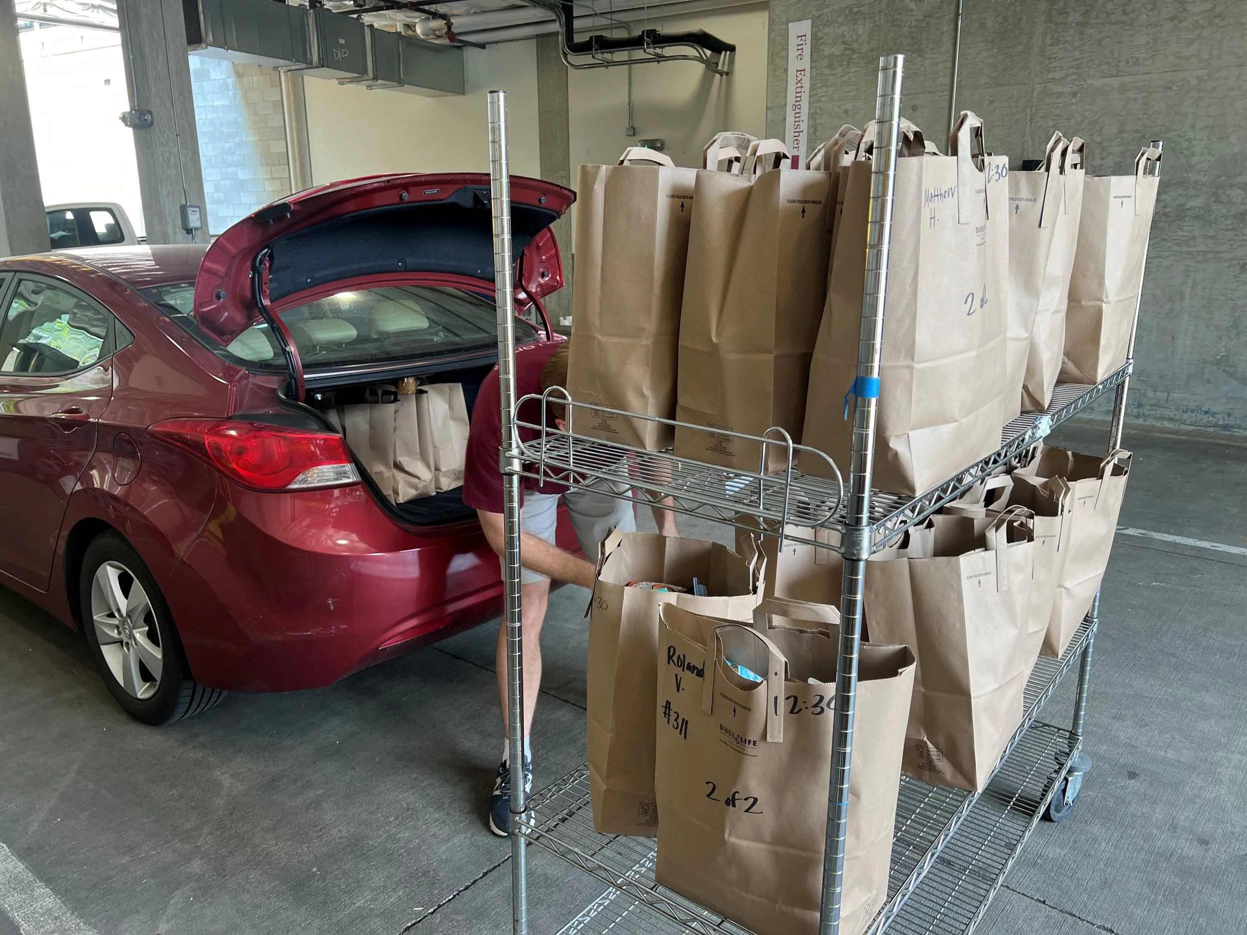 A red car is parked in a parking garage with its trunk open. Next to the car, a person is placing boxes on a metal cart filled with large brown paper bags, some labeled with names and numbers.
