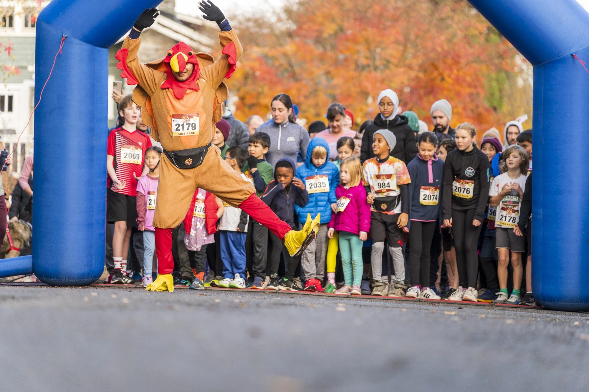 A person dressed in a turkey costume, complete with a red wattle and yellow beak, is crossing the finish line at a kids' race during autumn. The crowd of children and adults behind the person are watching and cheering. The scene is outdoors with fall foliage in the background.