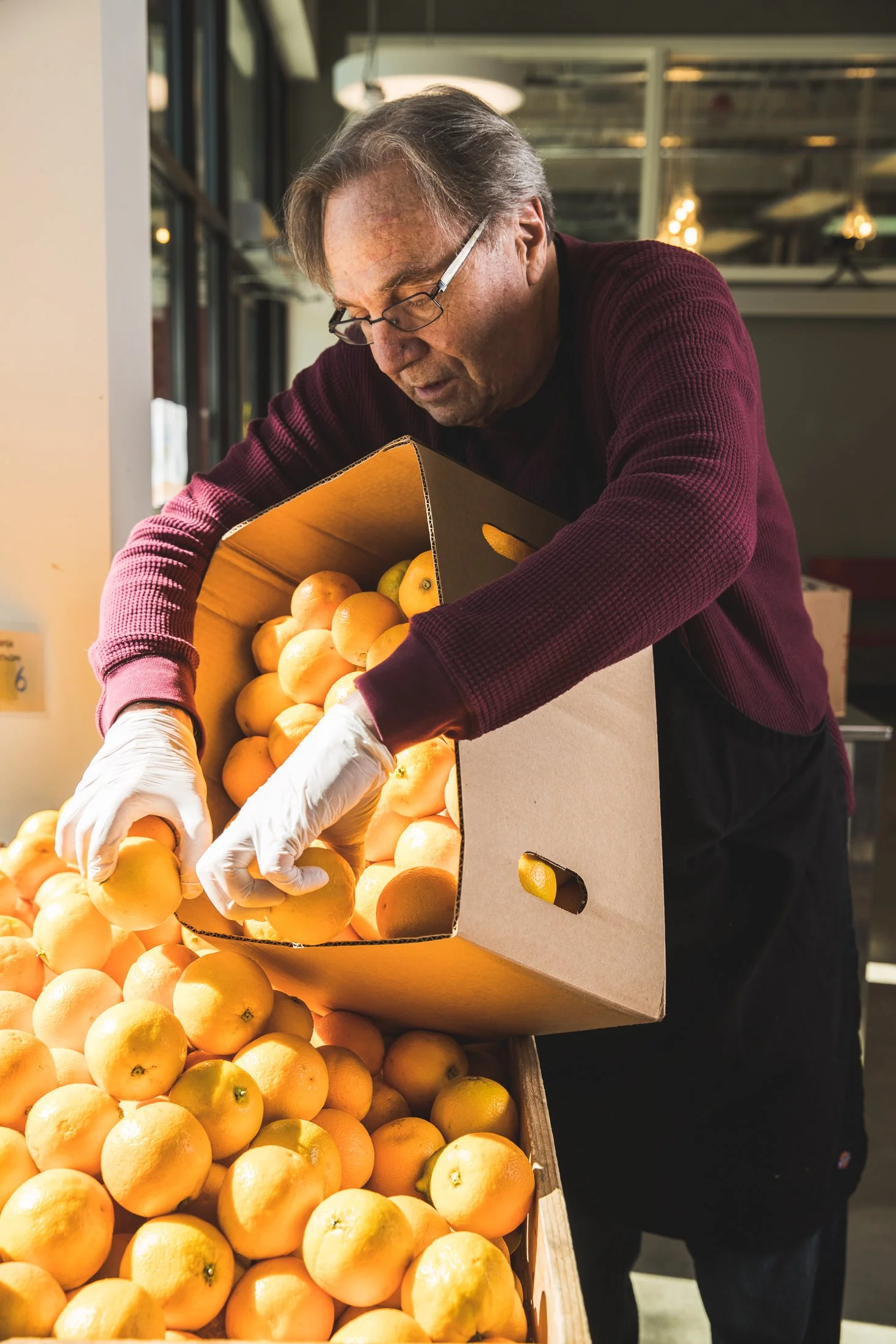 A man wearing glasses and a maroon sweater is placing yellow oranges from a cardboard box onto a display at a market.