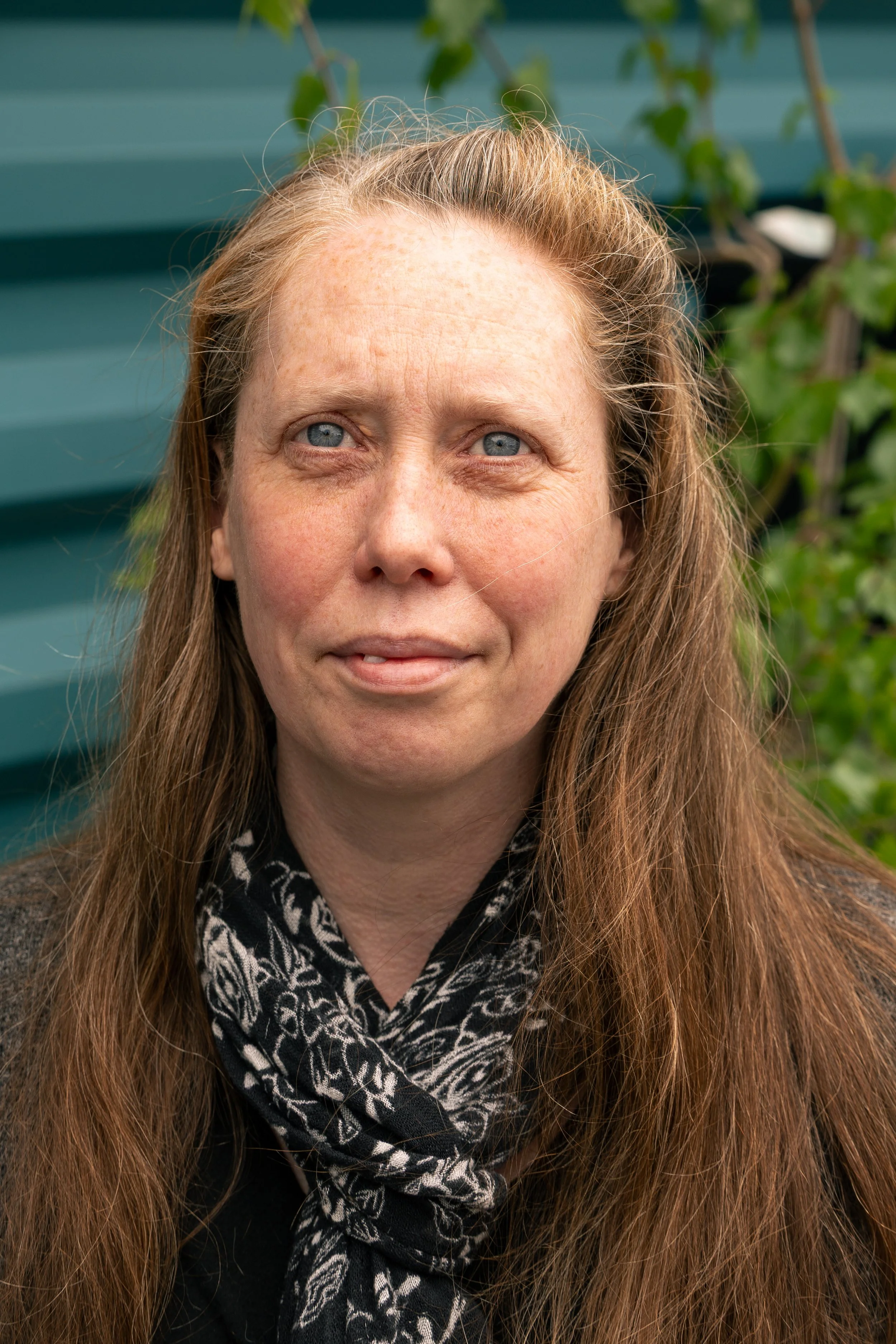 Close-up of a woman with long, reddish-brown hair and blue eyes, wearing a black top and a black-and-white patterned scarf, standing outdoors in front of a green fence with foliage.