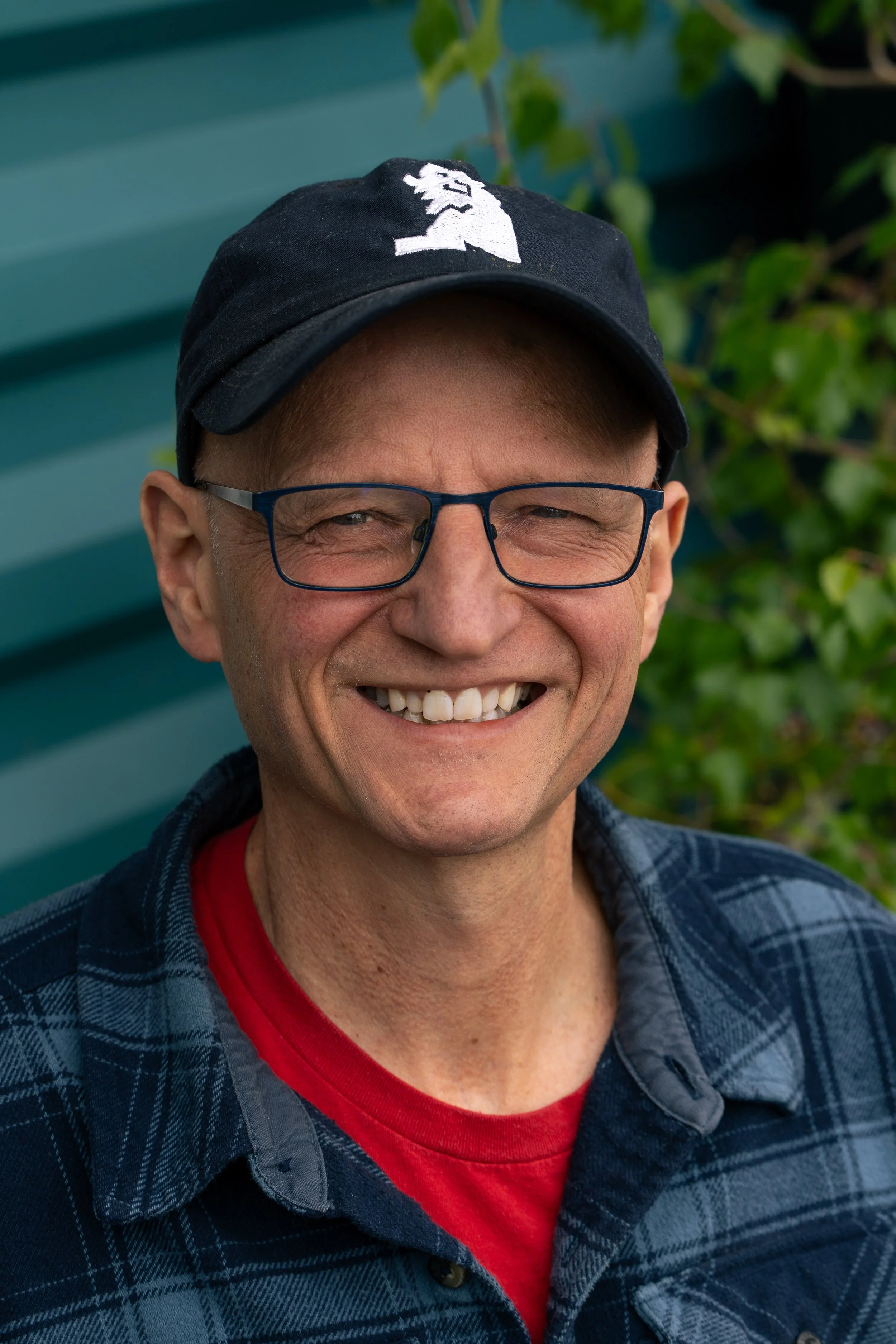 Close-up of a smiling man wearing glasses, a navy blue baseball cap with a white lion logo, a red shirt, and a blue plaid shirt, outside with green foliage and turquoise siding in the background.
