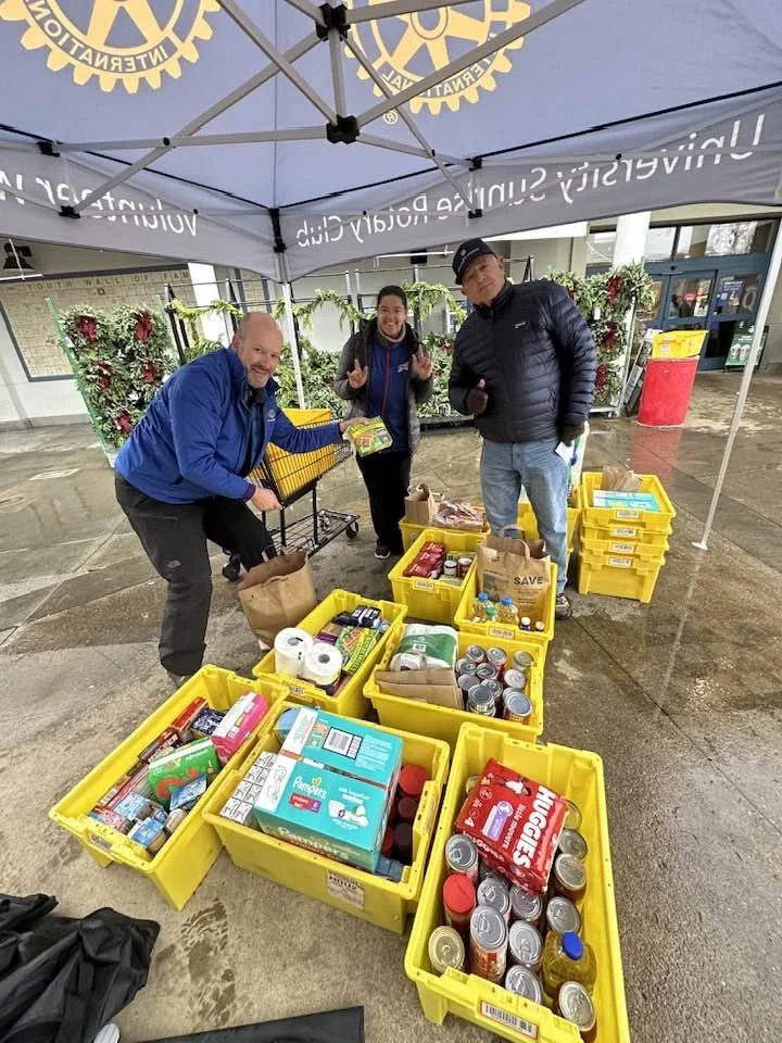 Three people standing under a Rotary International tent at a community event, organizing and sorting food donations in yellow plastic bins filled with canned goods and other non-perishable items.