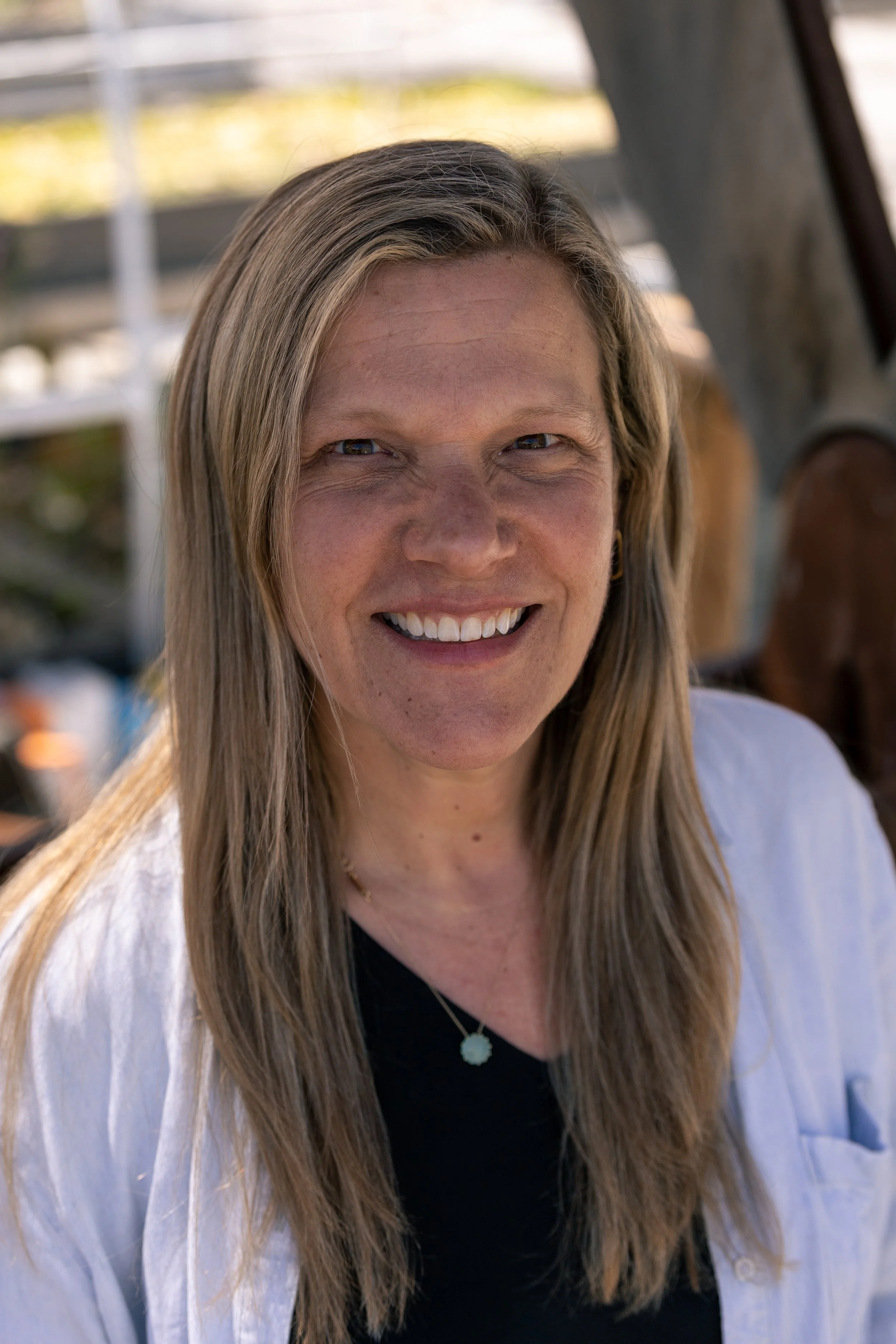Close-up of a woman with long blonde hair smiling outdoors, wearing a white blouse and black top, with a background of blurred outdoor structures.