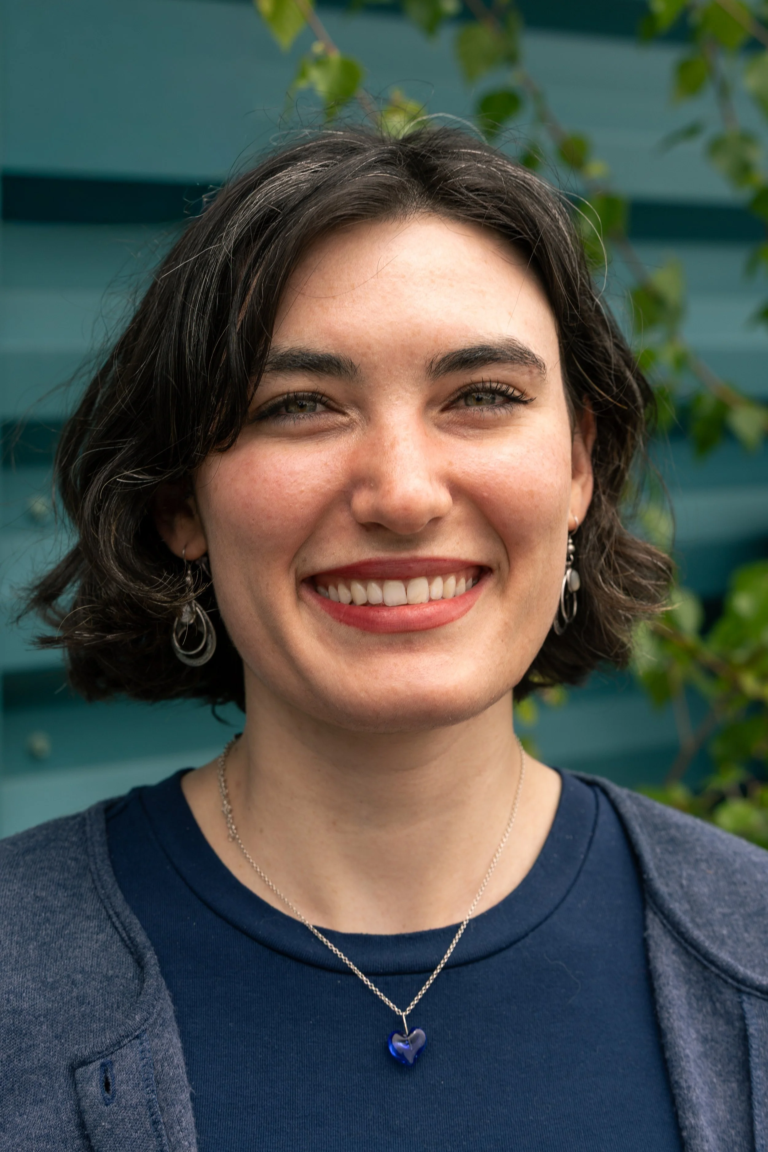 A smiling woman with short dark hair, wearing a navy blue shirt, a dark gray cardigan, and a silver necklace with a blue heart-shaped pendant. She is outdoors with green leaves and blue siding in the background.