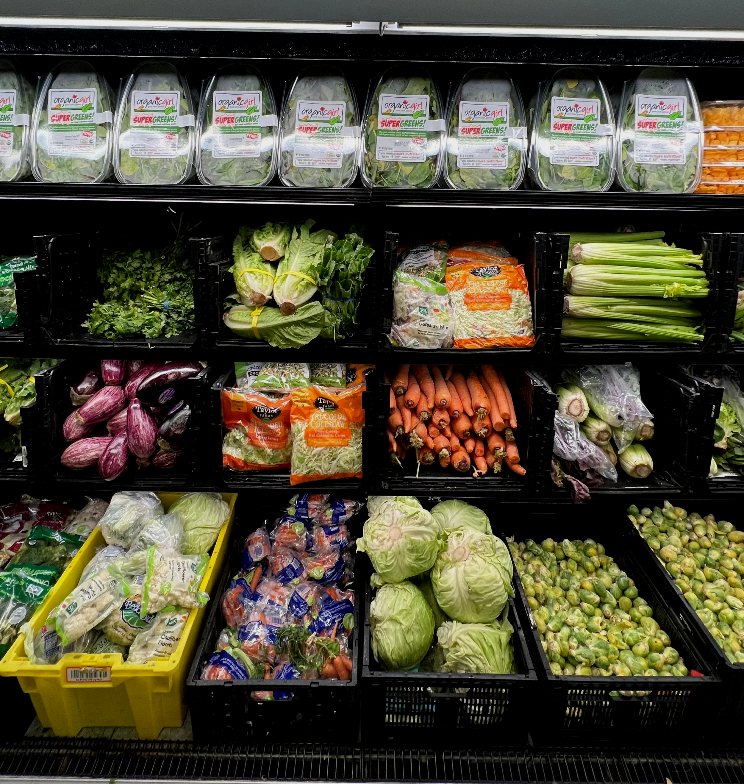 Multiple shelves of fresh vegetables including leafy greens, romaine lettuce, carrots, celery, and purple eggplants in a grocery store produce section.