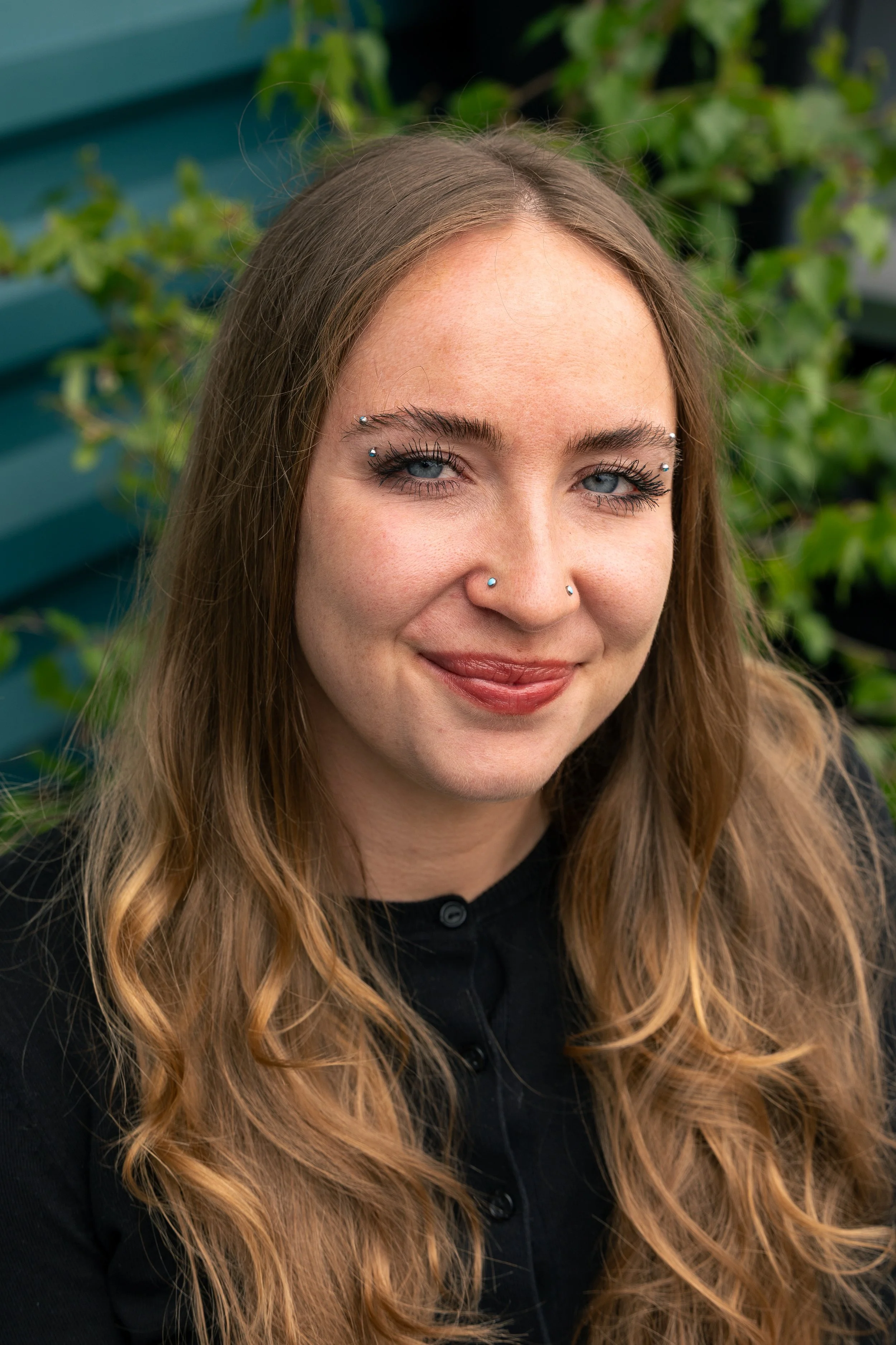 Close-up of a young woman with long, wavy, light brown hair, wearing piercings near her eyebrows and nose, smiling softly, outdoor background with green foliage and a teal wooden fence.
