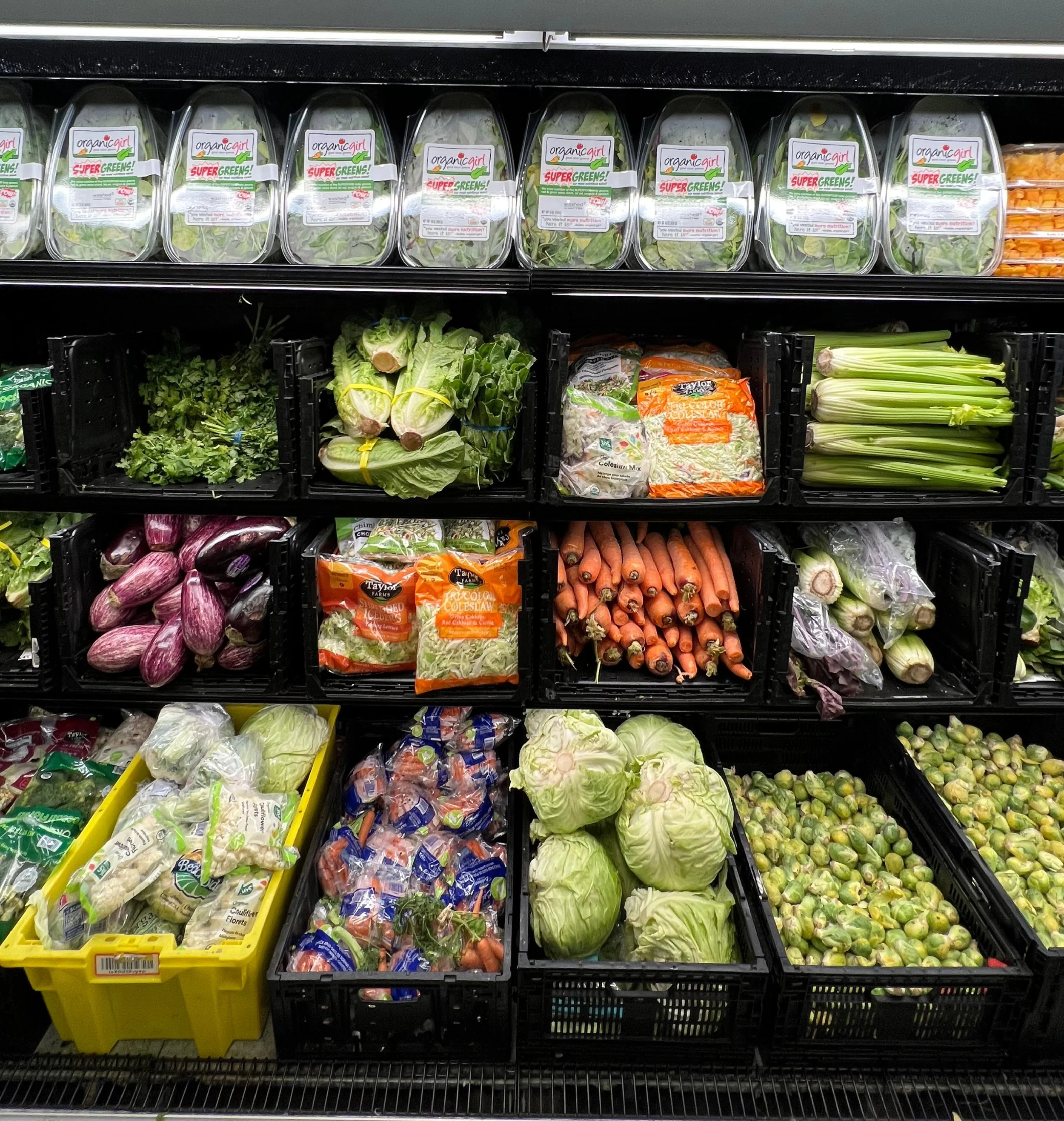 Fresh vegetables on store shelves including eggplants, lettuce, carrots, celery, and spinach.