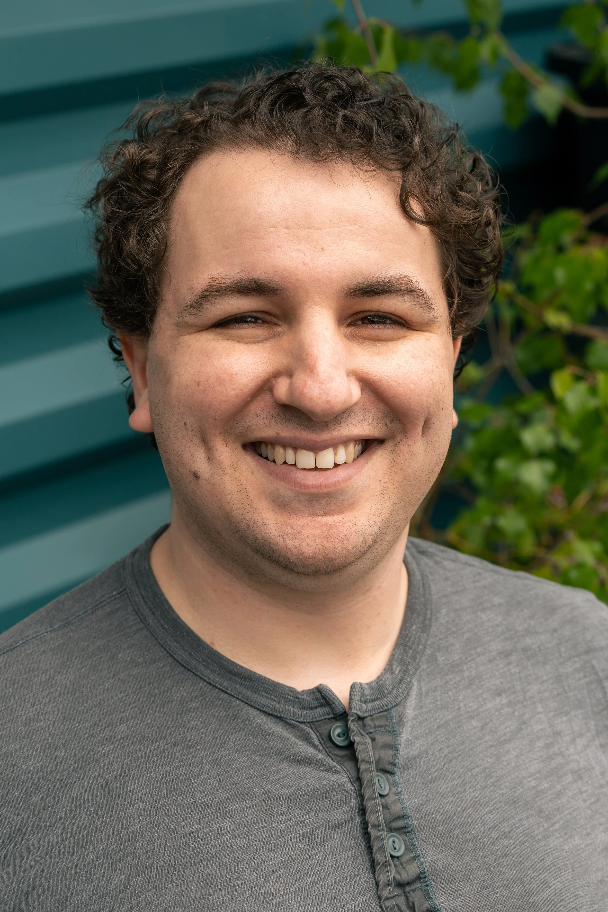 A close-up photo of a man with curly dark hair, smiling, wearing a gray shirt with a buttoned collar, standing outdoors with a blue-green background and some green leaves.