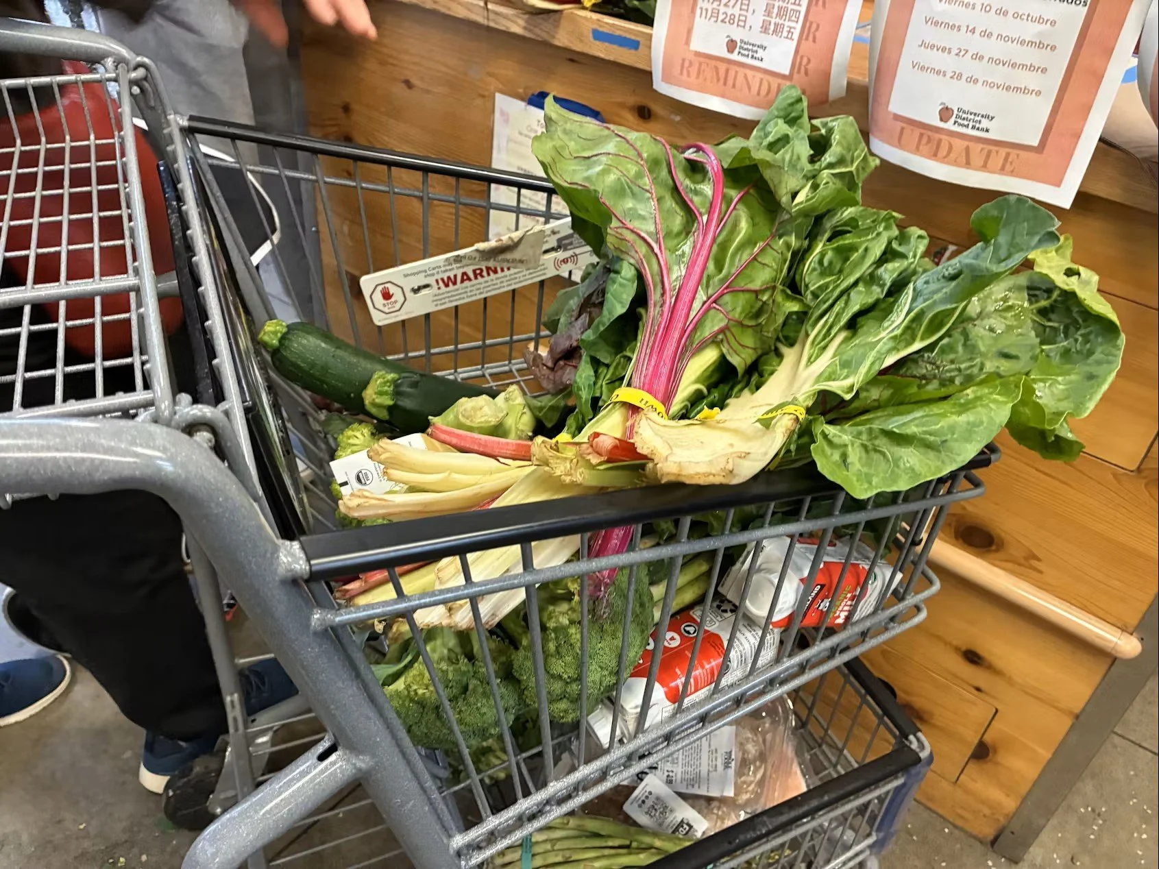 A shopping cart filled with fresh vegetables including zucchini, broccoli, and Swiss chard at a grocery store. A wooden surface with signs and other shopping items is visible in the background.