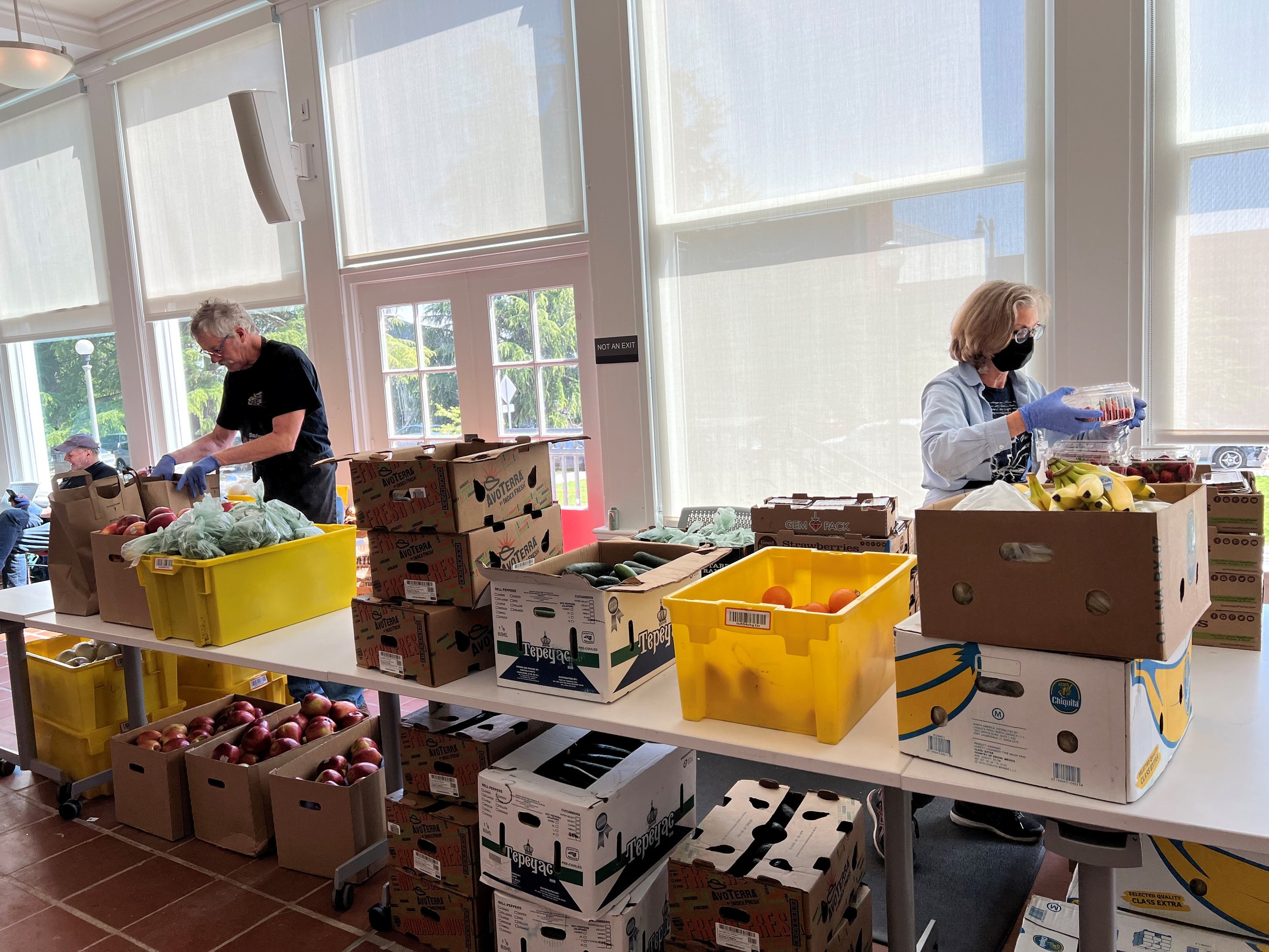 People sorting and packing fresh produce, including apples, bananas, and other fruits, in a bright room with large windows.