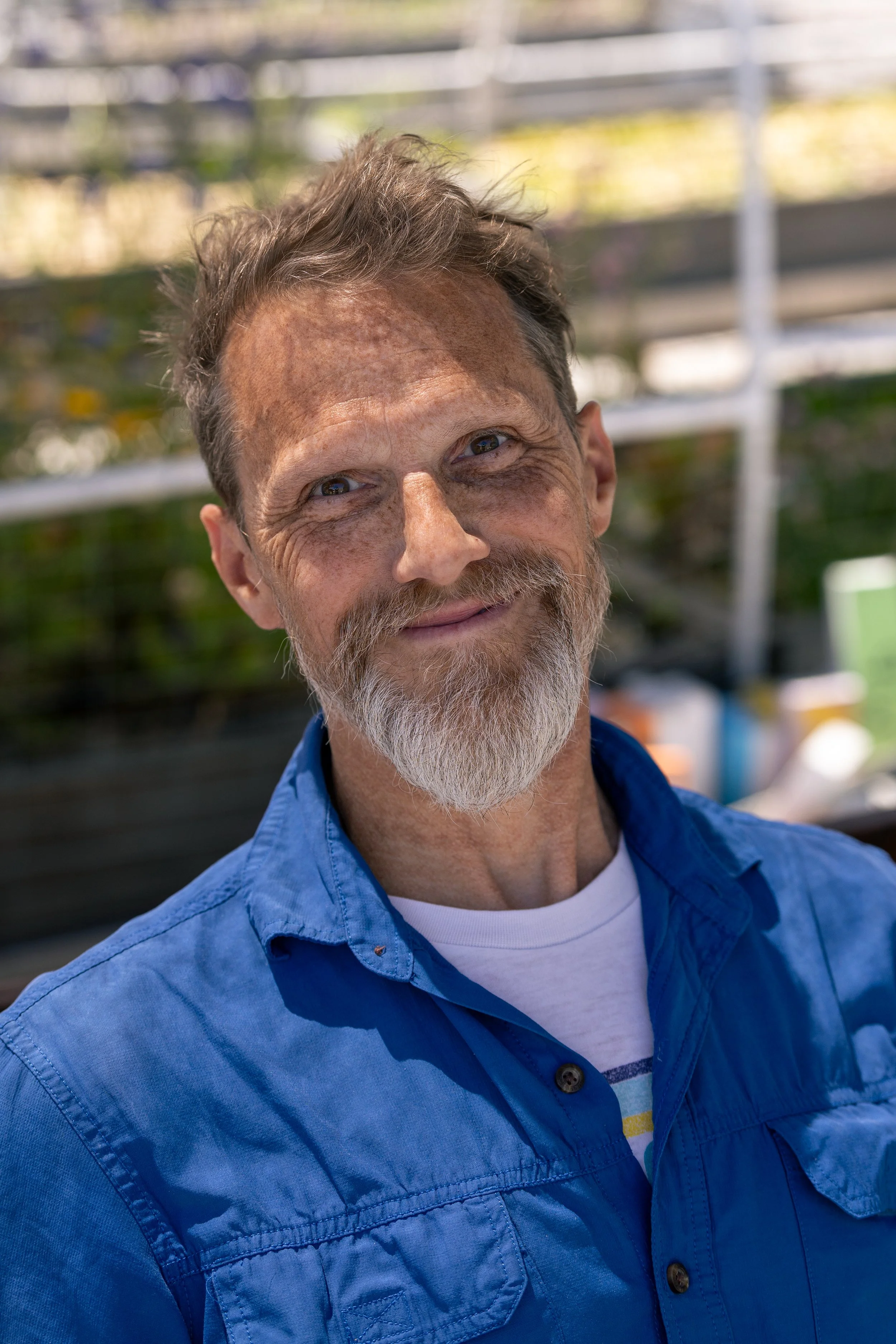 Close-up of a smiling middle-aged man with gray hair and beard wearing a blue shirt outdoors with blurred background.