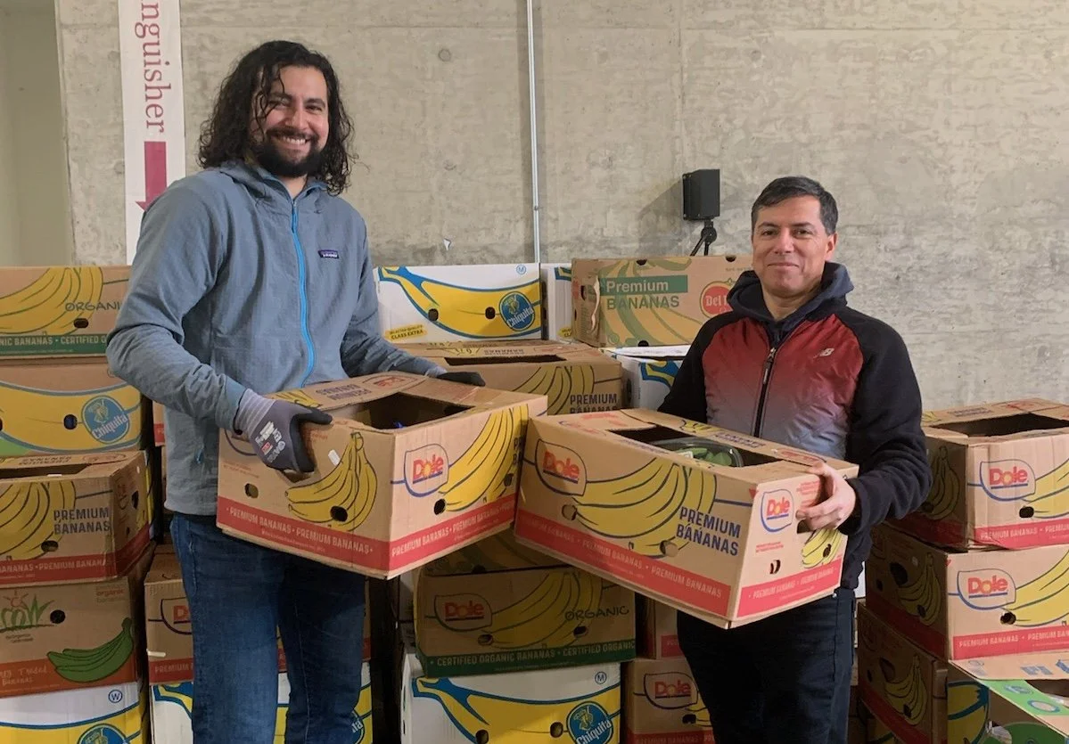 Two men standing inside a warehouse surrounded by boxes of bananas. The man on the left has curly hair, a beard, and is wearing a gray hoodie. The man on the right has short dark hair and is wearing a black and red jacket. They are holding boxes of bananas, and there are more boxes stacked around them.