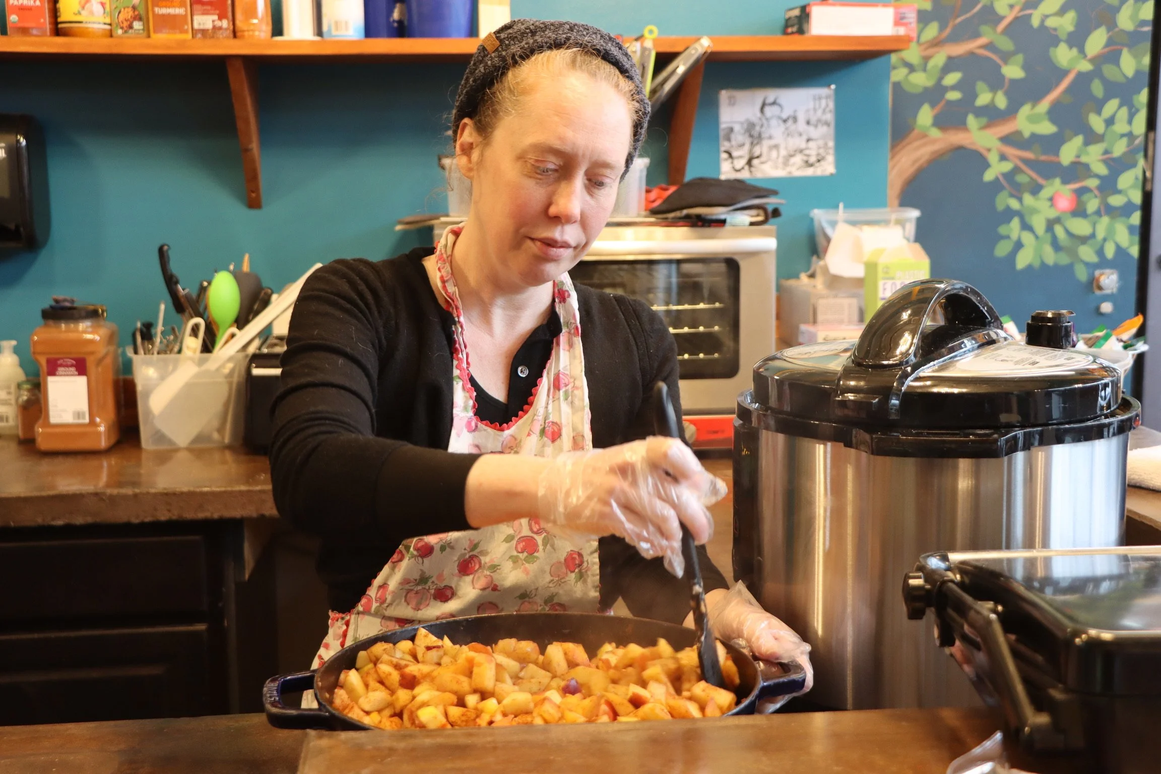 A woman in a floral apron and a headband uses tongs to place roasted potatoes into a container in a crowded kitchen or restaurant setting.