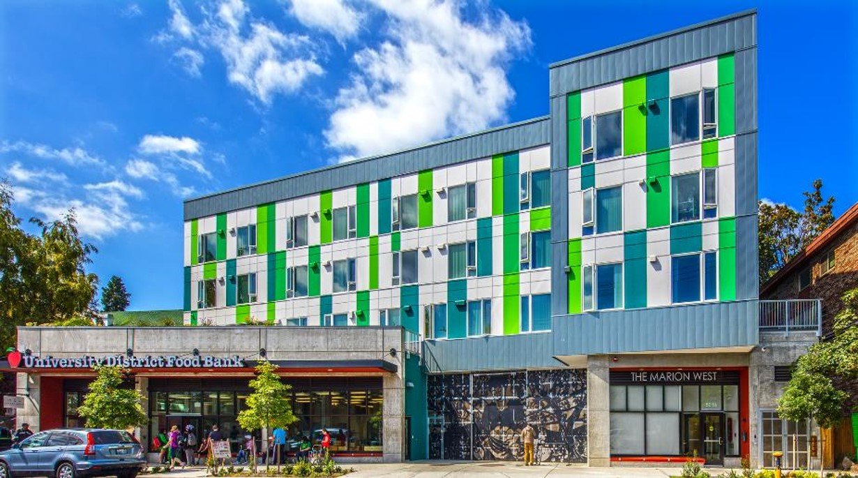 Modern multi-story building with green and white exterior panels, adjacent to a storefront with a sign for University District Food Bank, and people walking outside on a sunny day.