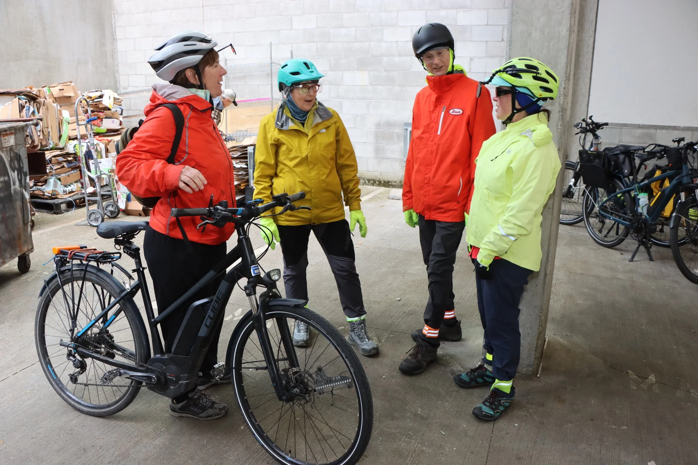 Four people wearing helmets and outdoor jackets engaged in conversation next to bicycles in an alley with concrete walls and a pile of cardboard boxes.