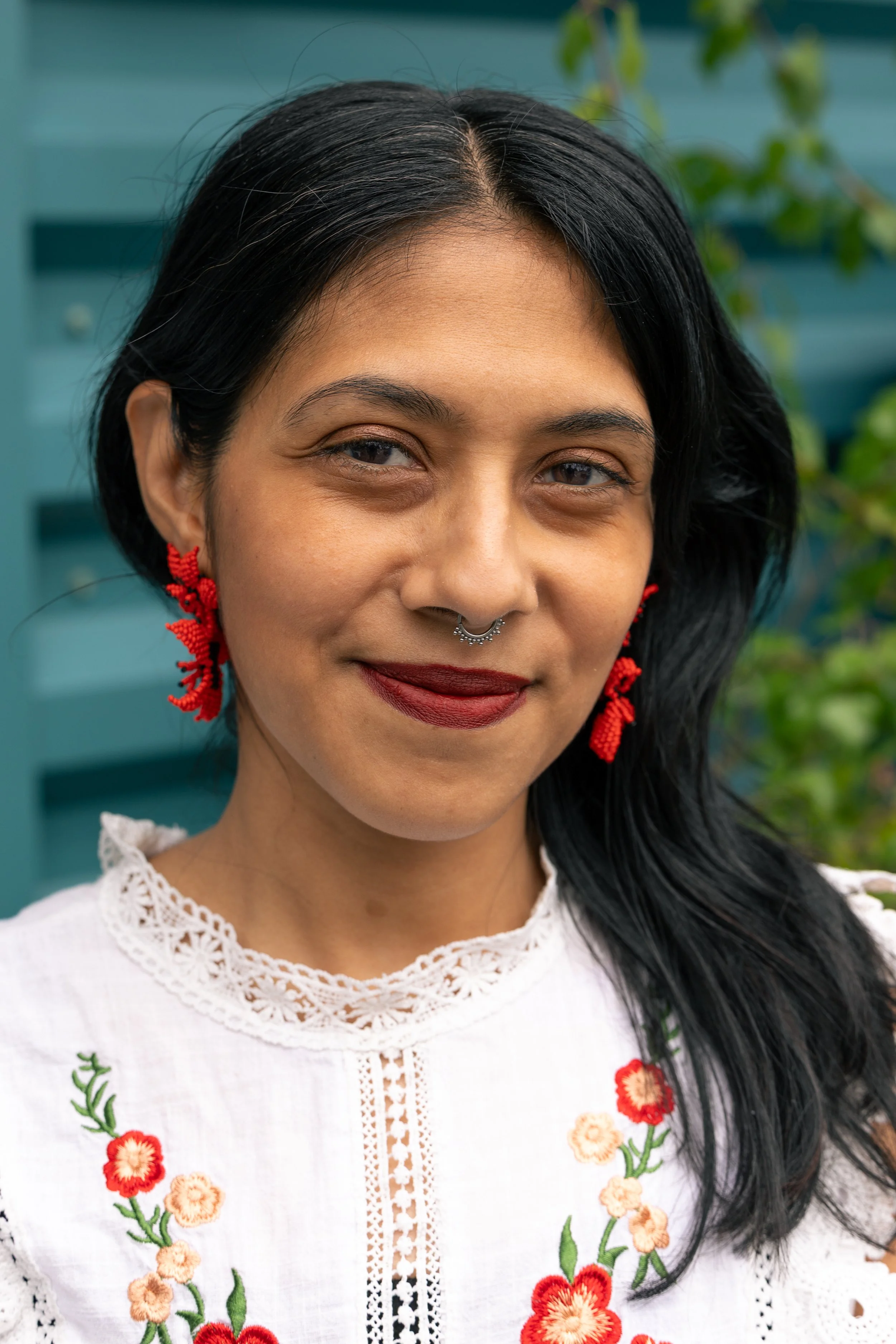 Close-up of a woman with dark hair, wearing red beaded earrings, a silver septum piercing, and a white embroidered blouse, smiling outdoors with a blue building and greenery in the background.