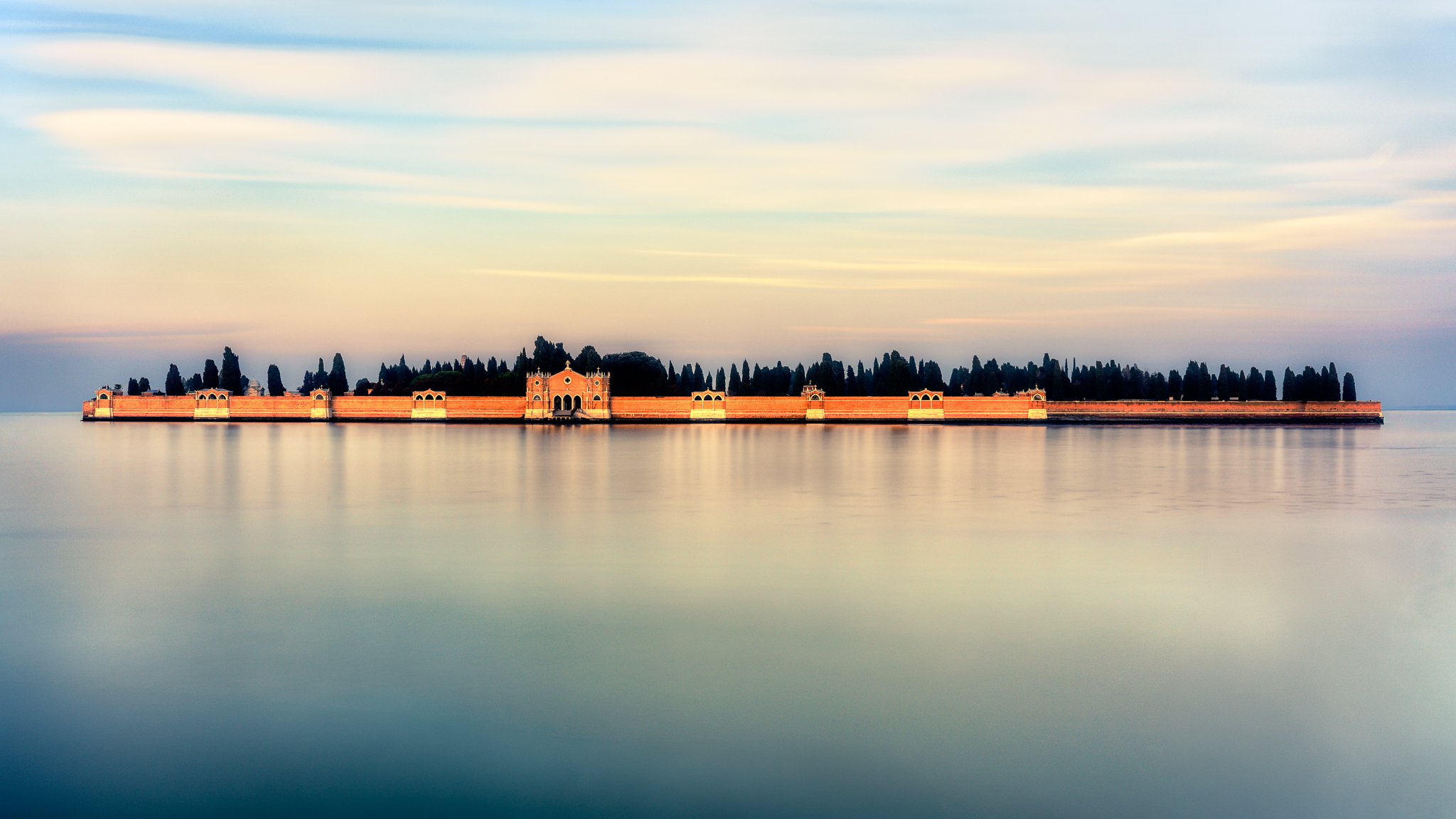 San Michele island in Venice at twilight with glowing brick walls, cypress trees, and calm lagoon under a pastel sky.