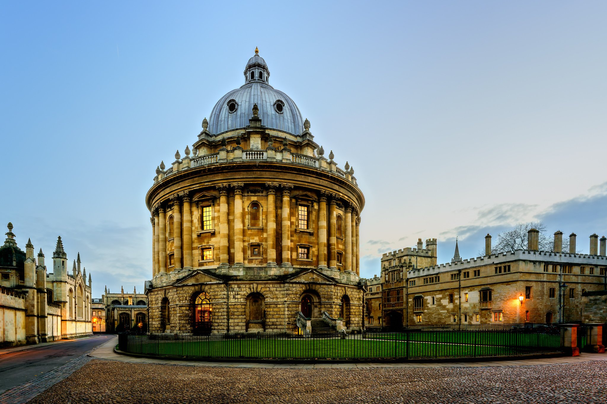 Radcliffe Camera library in Oxford illuminated at dusk with historic university buildings and cobbled street in the foreground.