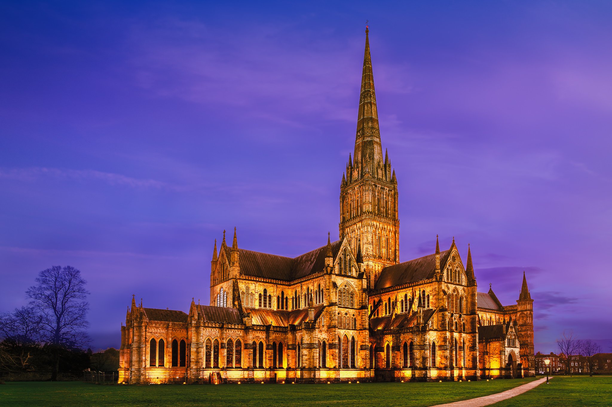 Salisbury Cathedral illuminated at blue hour with warm lighting against a deep blue sky and surrounding lawn.