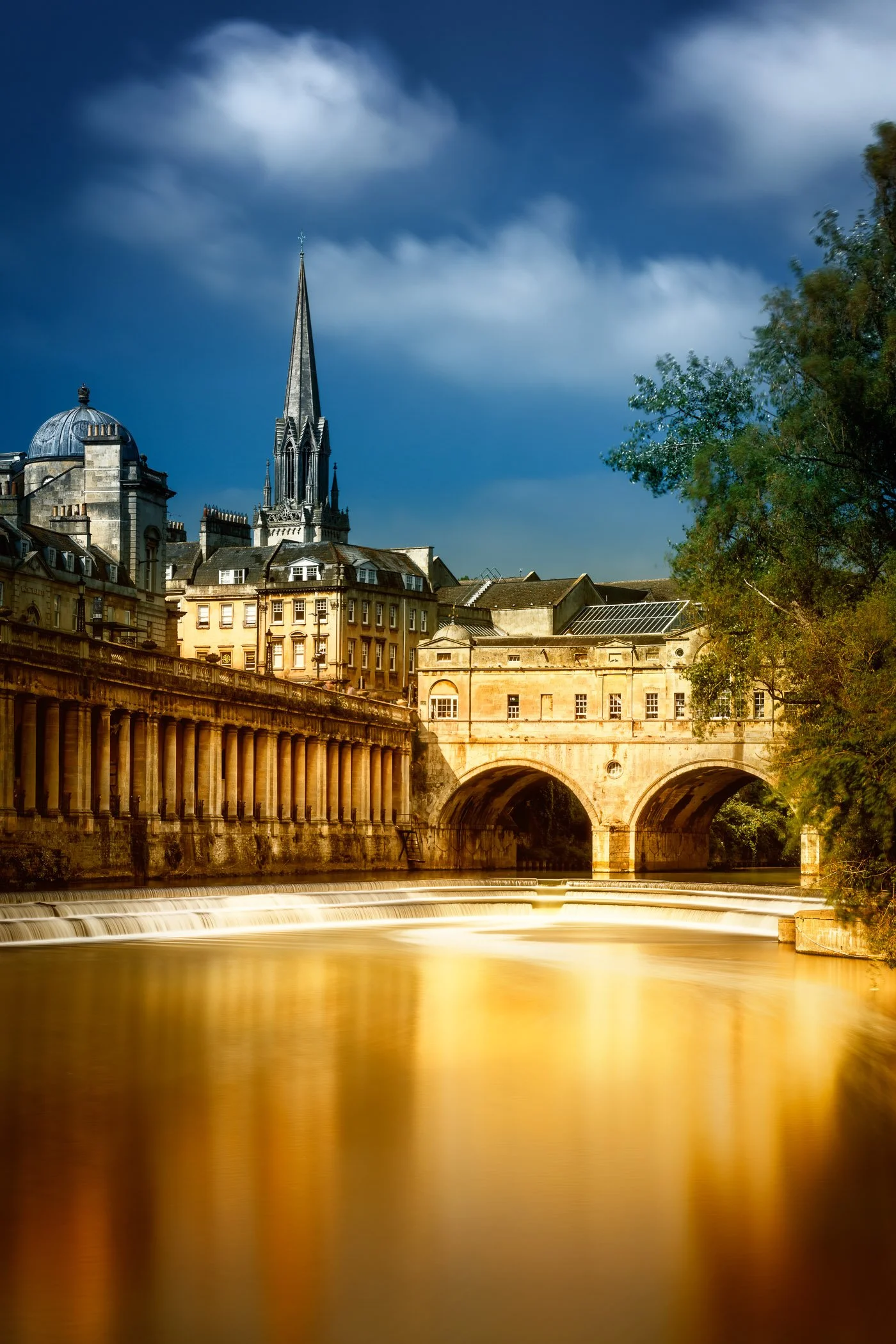 Pulteney Bridge in Summer A daytime perspective of Pulteney Bridge surrounded by lush summer greenery along the riverbanks.