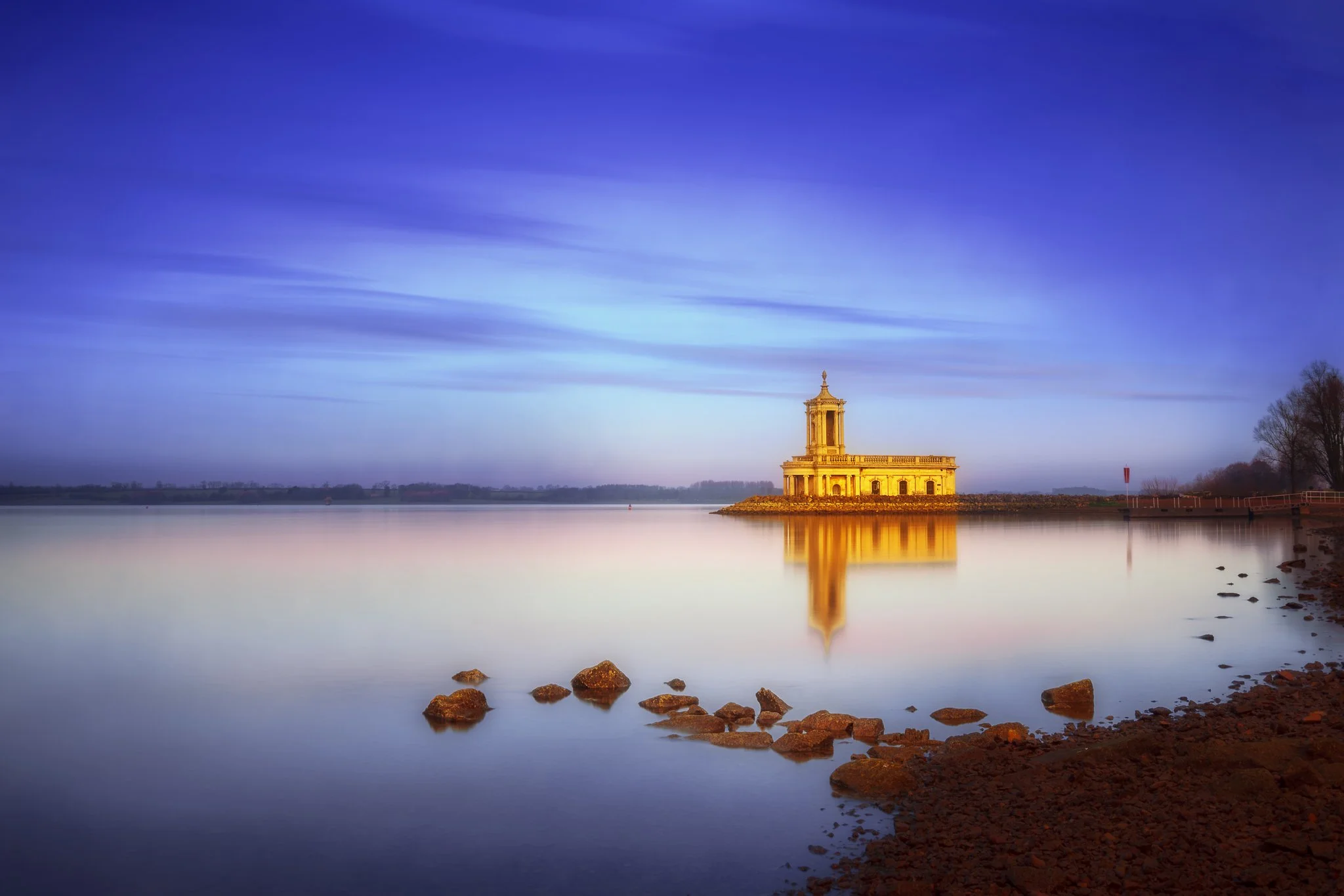 A historic church called Normanton  by a calm body of water, with a clear sky and some clouds, trees on the right side, and rocks in the foreground.