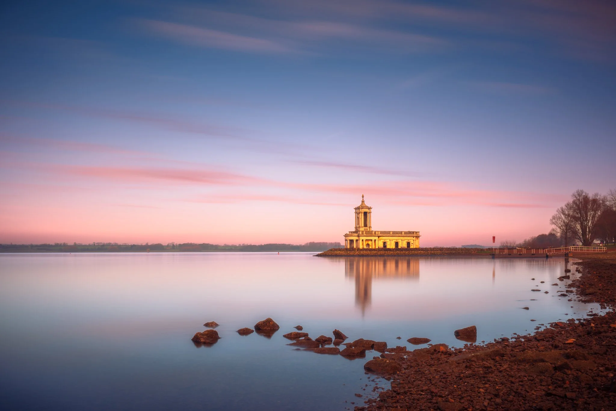 Scenic view of Normanton Church on a small island in a calm body of water during sunset, with a colourful sky and bare trees on the right side.