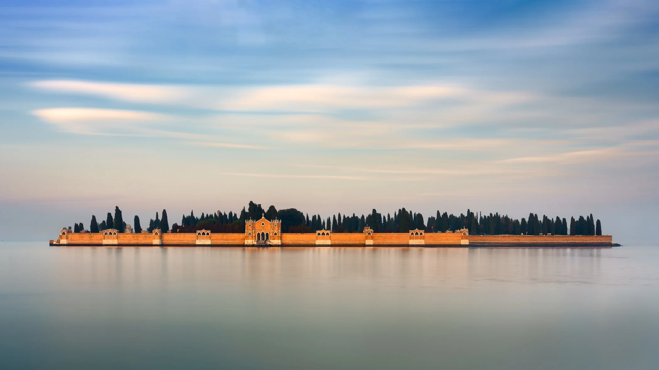 Long-exposure photograph of San Michele island in Venice, with terracotta walls and dark cypress trees beneath a soft blue and peach sky reflected in calm lagoon water.