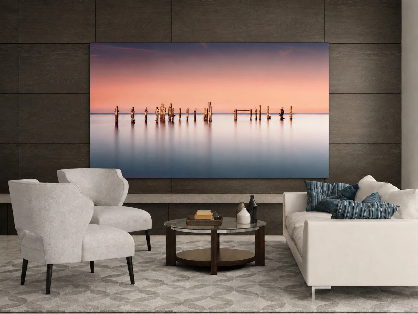 Modern living room with white armchairs, a white sofa with striped pillows, a glass coffee table with books and vases, dark wood wall panels, a large landscape wall art of a sunset over water with wooden posts, and patterned carpet.