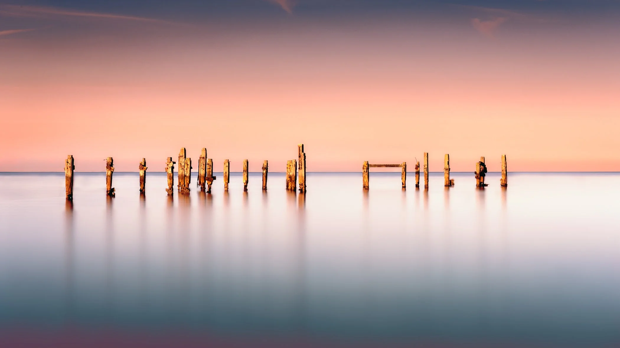 Echoes of Time A serene minimalist long exposure photograph capturing weathered wooden sea posts standing peacefully against pastel horizon tones.