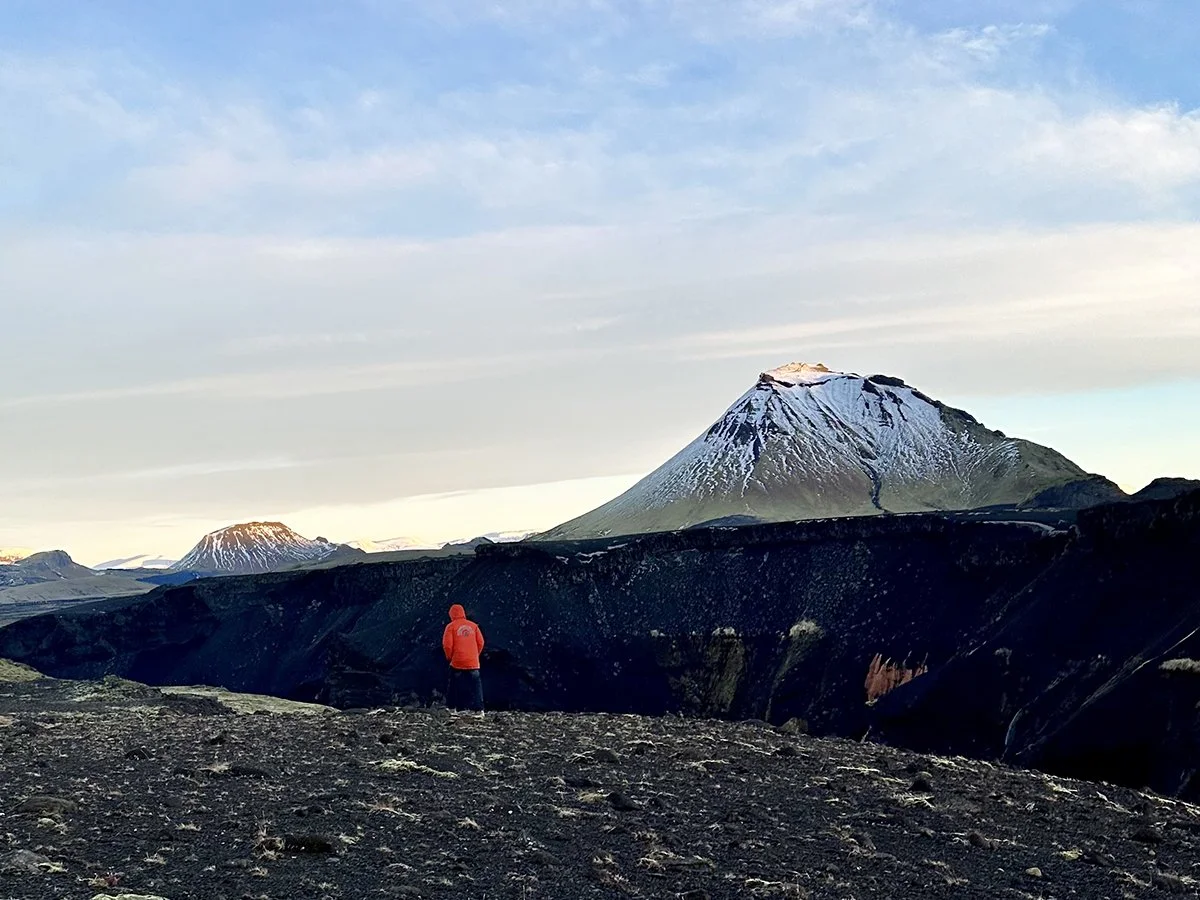 Traveler standing on Iceland’s volcanic highlands overlooking a snow-covered cone mountain.