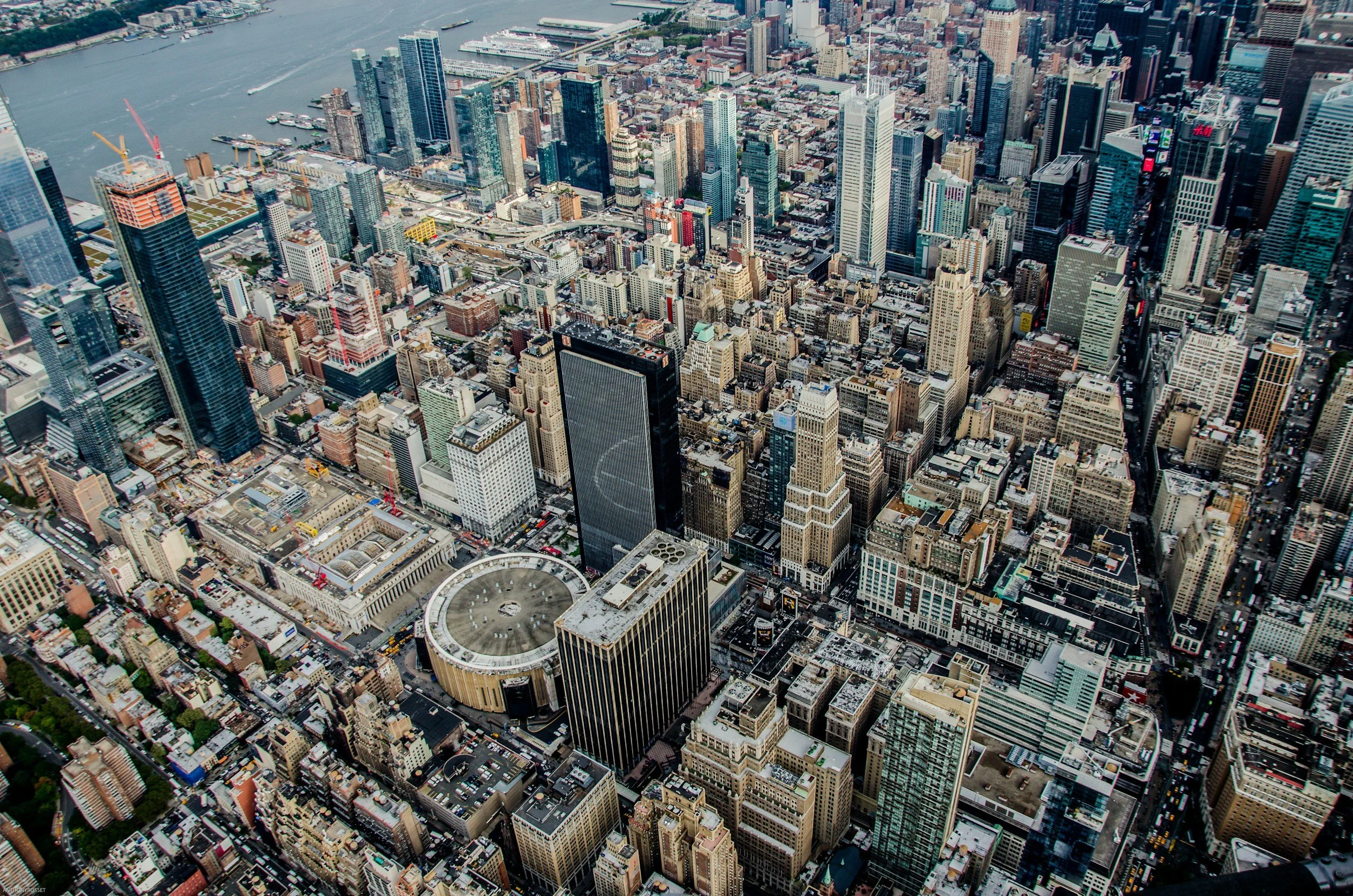 Aerial view of New York City skyline showing numerous tall skyscrapers, streets, and a river in the background.