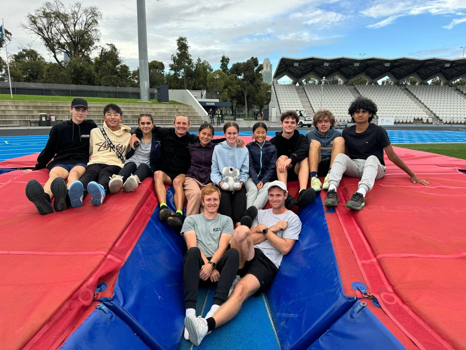 A group of diverse young athletes and coaches sitting on a red and blue high jump mat at an outdoor stadium with a running track and stadium seats in the background.