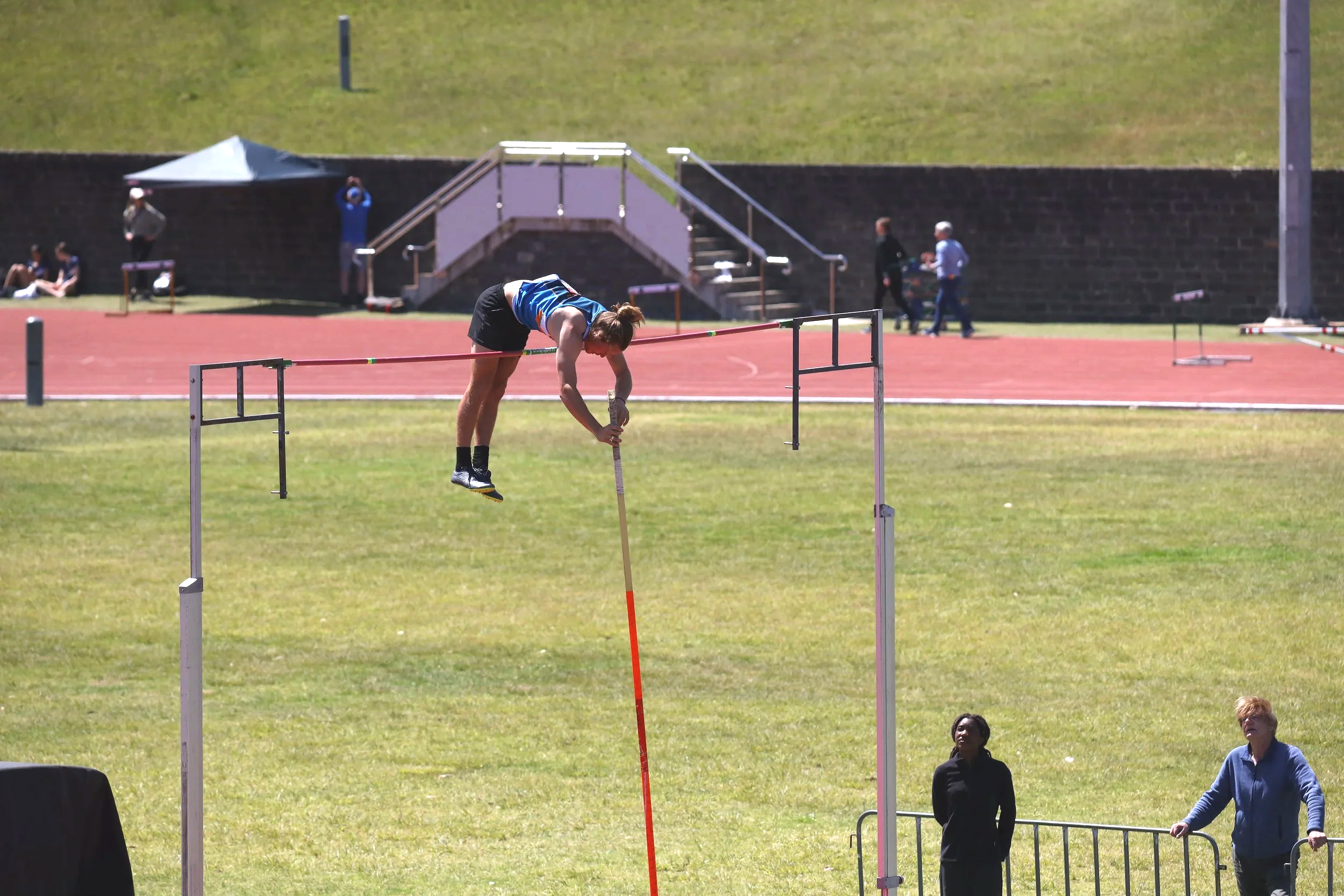 A female pole vaulter bends down on a track, preparing with a pole, while a few spectators watch nearby. The setting is an outdoor athletic field with grass and a running track in the background.