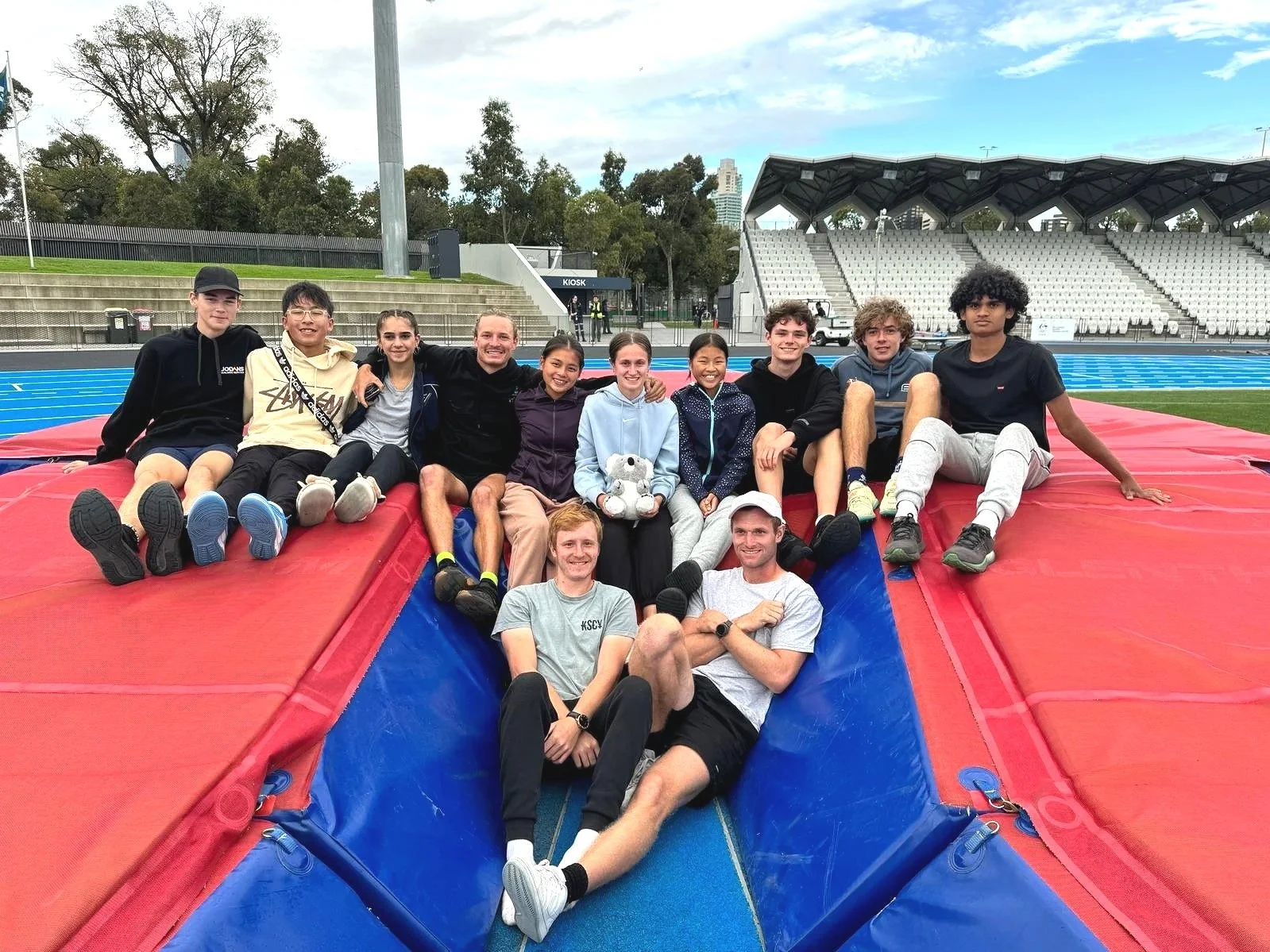 A group of young athletes and coaches sitting and lying on a red and blue high jump mat at a stadium track and field area, with empty bleachers in the background.